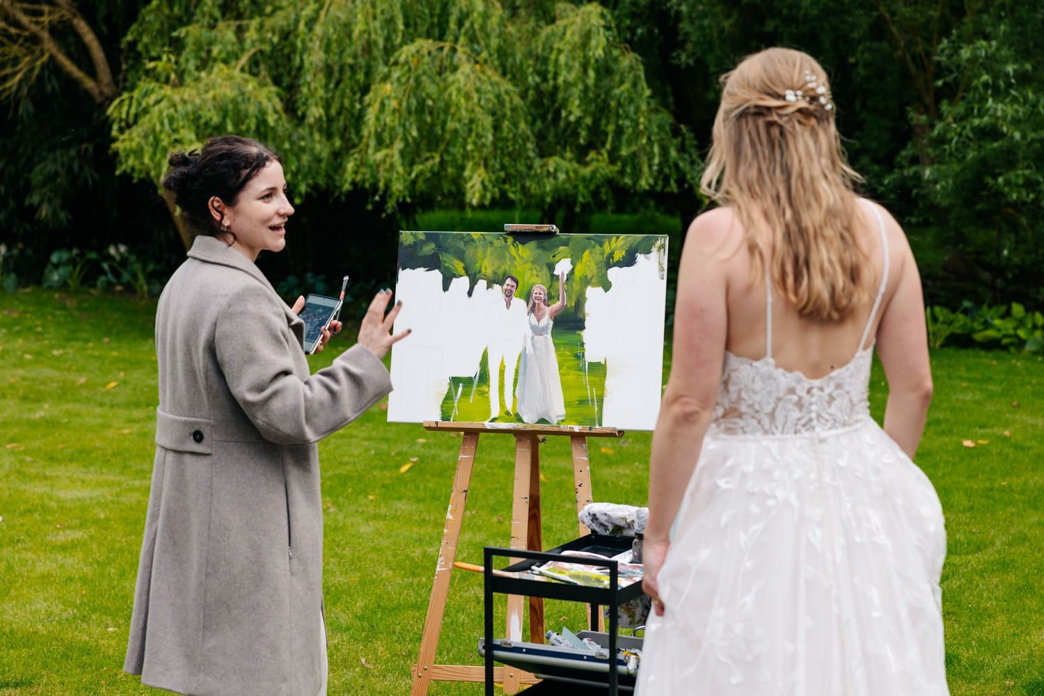 Live wedding artist showing canvas painting to bride in white gown in garden.