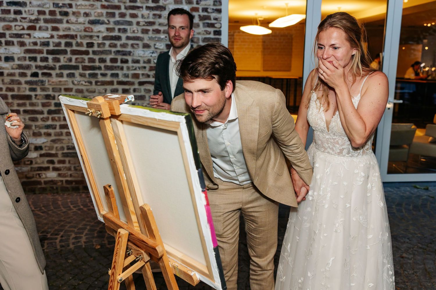 Surprised bride covers mouth as groom reveals live painting artwork at evening wedding reception.