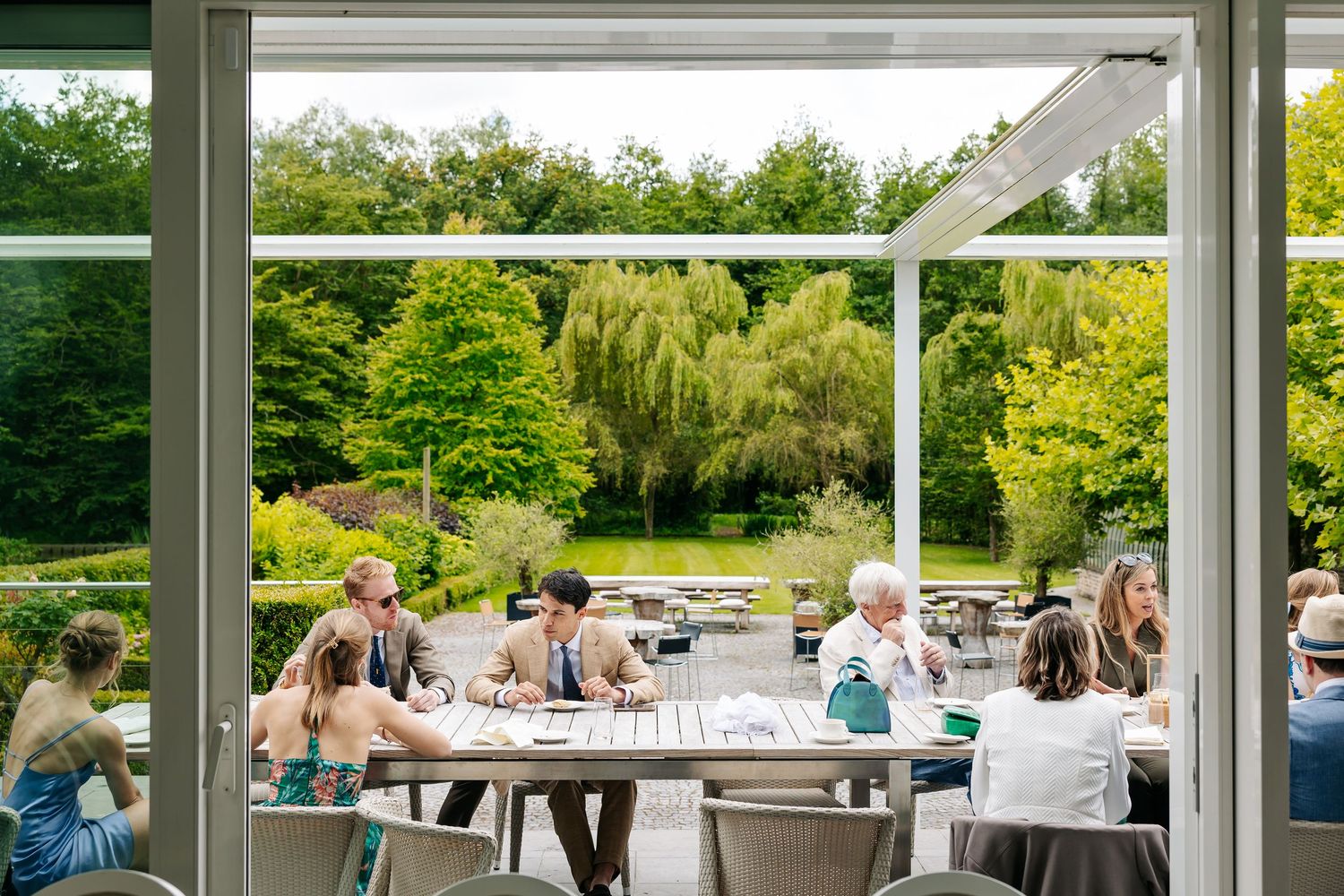 Wedding guests dining inside a modern glass conservatory overlooking a lush green garden landscape.