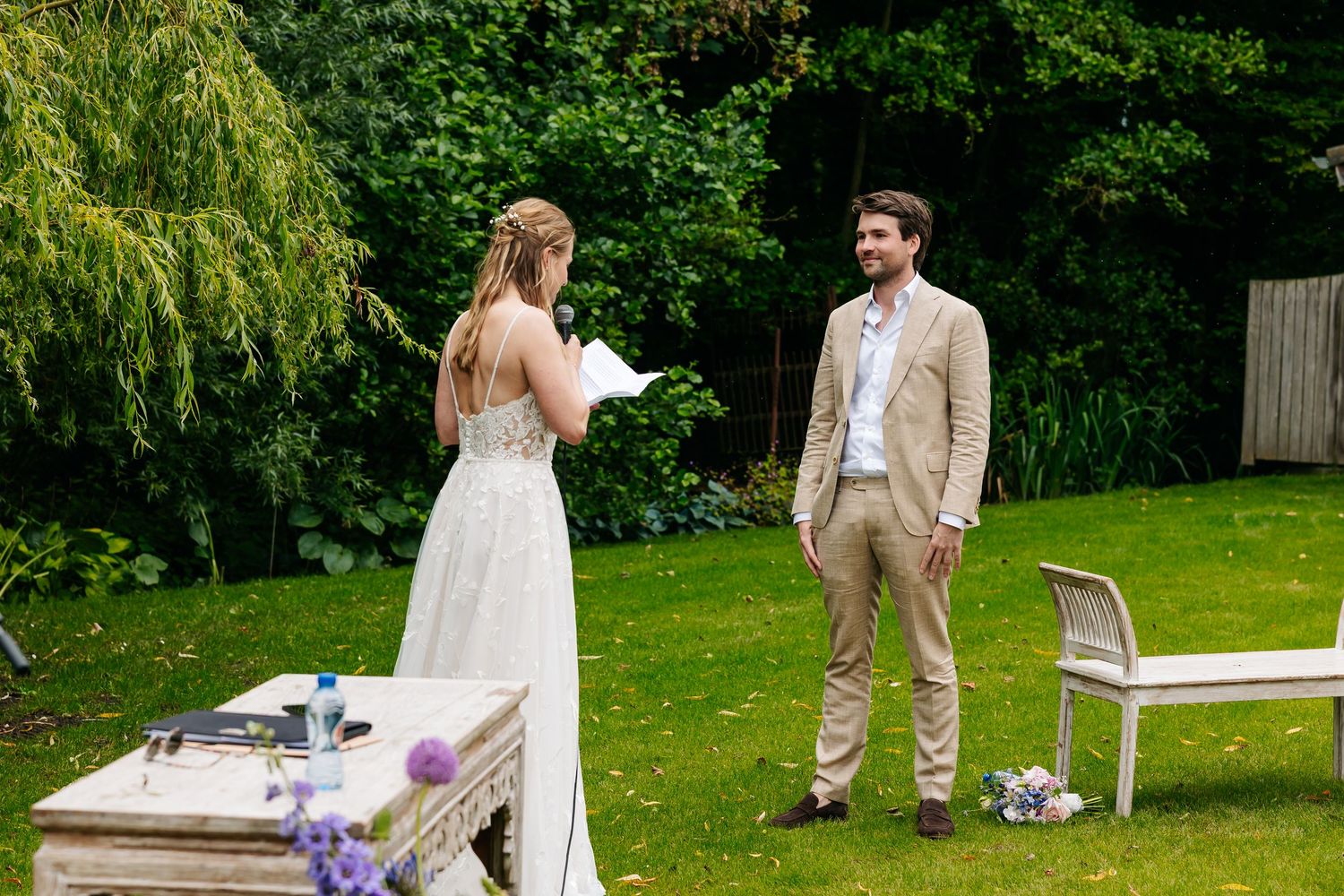 Bride reading vows to groom during an outdoor garden wedding ceremony on a sunny day.