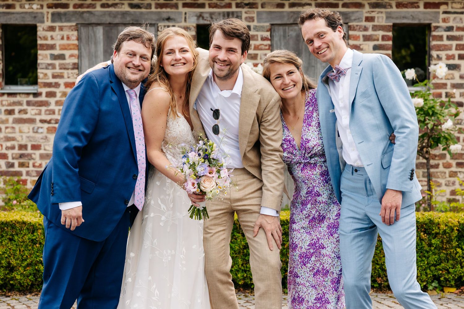 Bride and groom in beige suit smiling with three guests in colorful outfits at outdoor wedding.