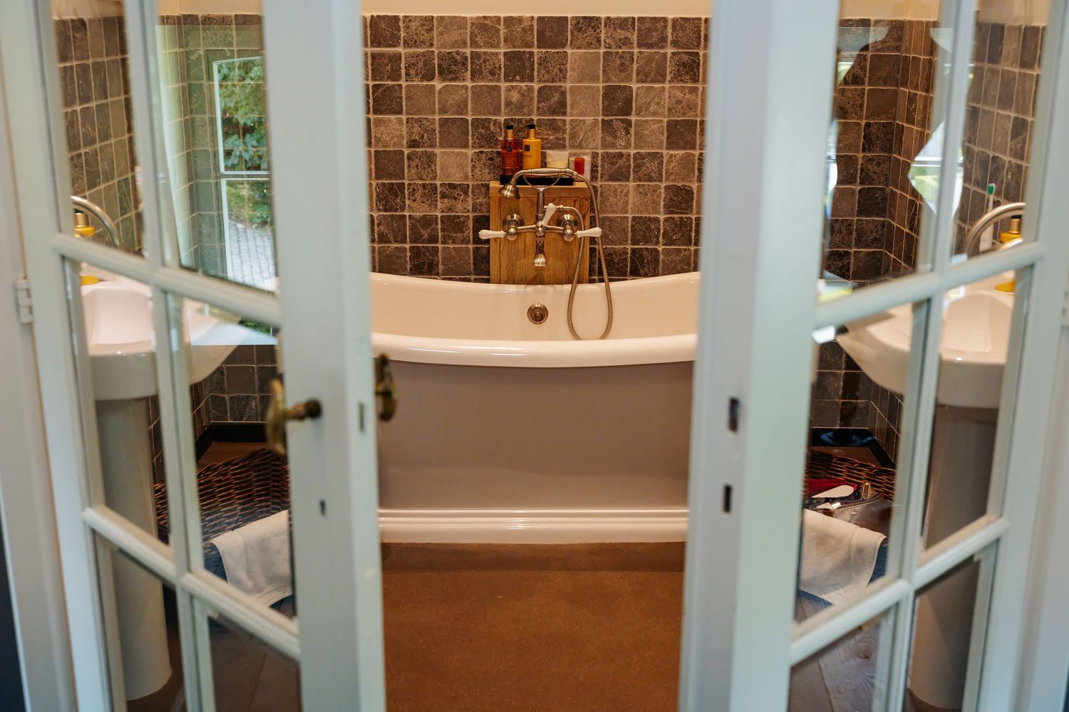 Elegant bathroom with freestanding bathtub viewed through open white French doors with gold fixtures.