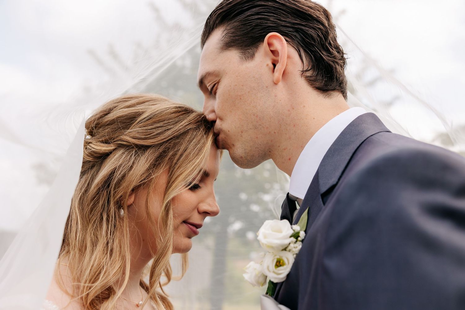 Groom gently kisses the forehead of his smiling bride beneath a sheer veil on their wedding day.