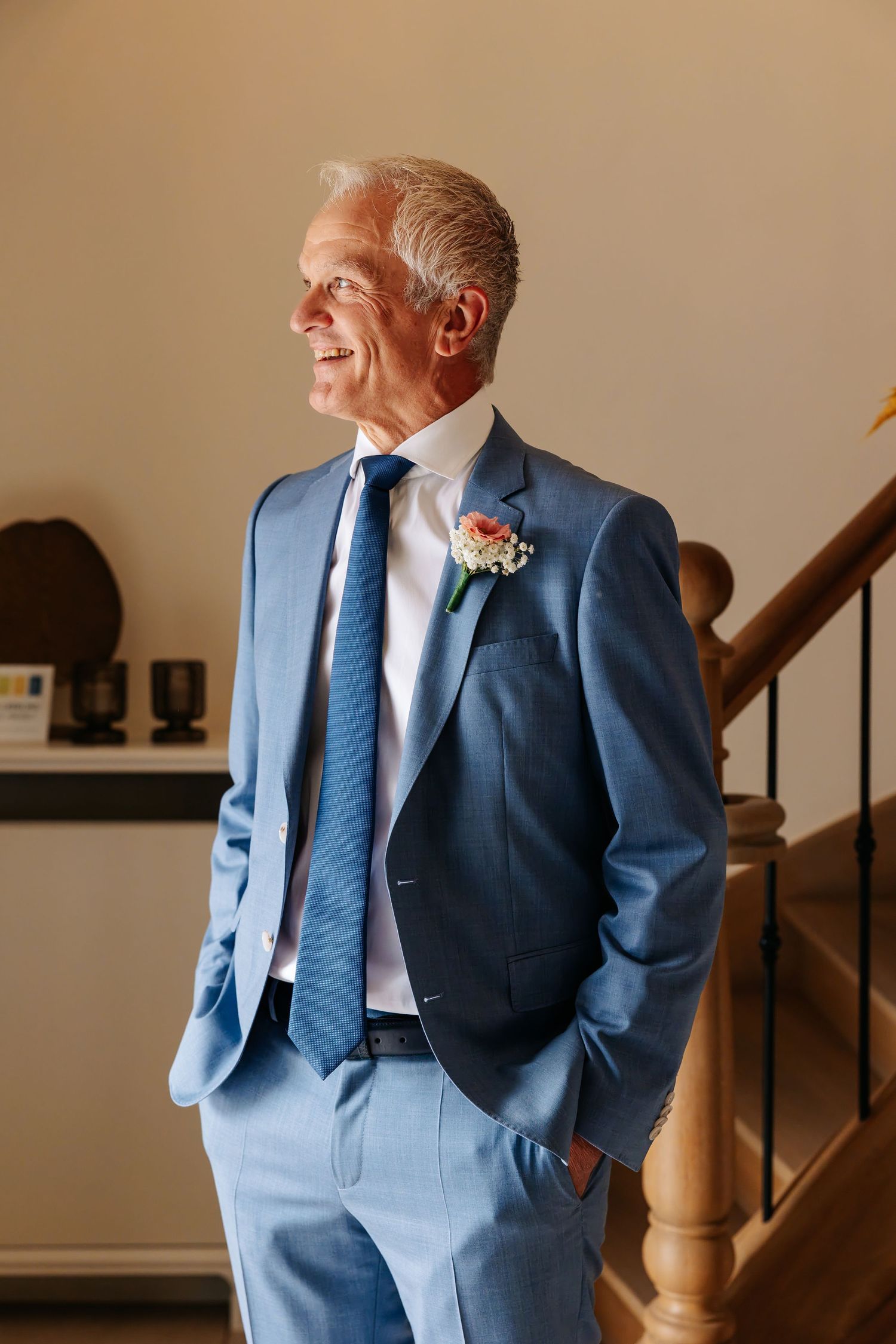 Stylish groom in blue suit with boutonniere gazes sideways while standing near a wooden staircase.