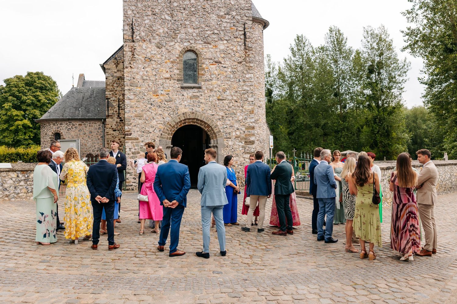 Wedding party and guests gather on cobblestone courtyard outside rustic stone church.