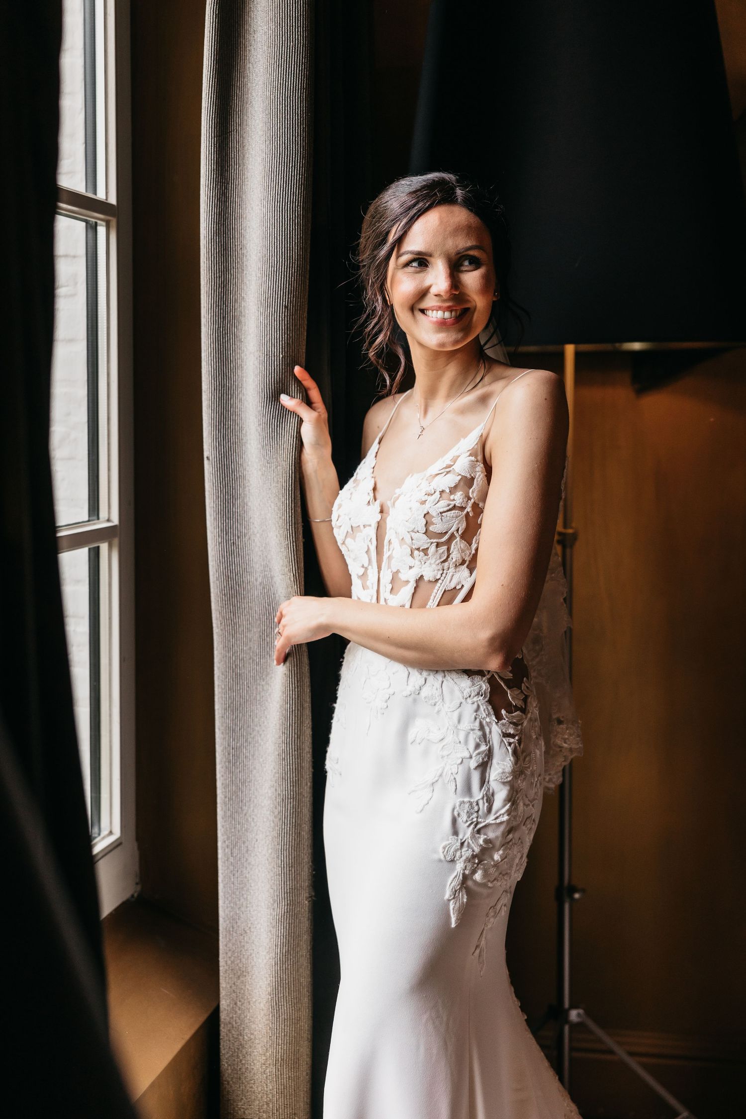 Elegant bride in fitted lace wedding dress smiles beautifully while posing beside a curtained window.