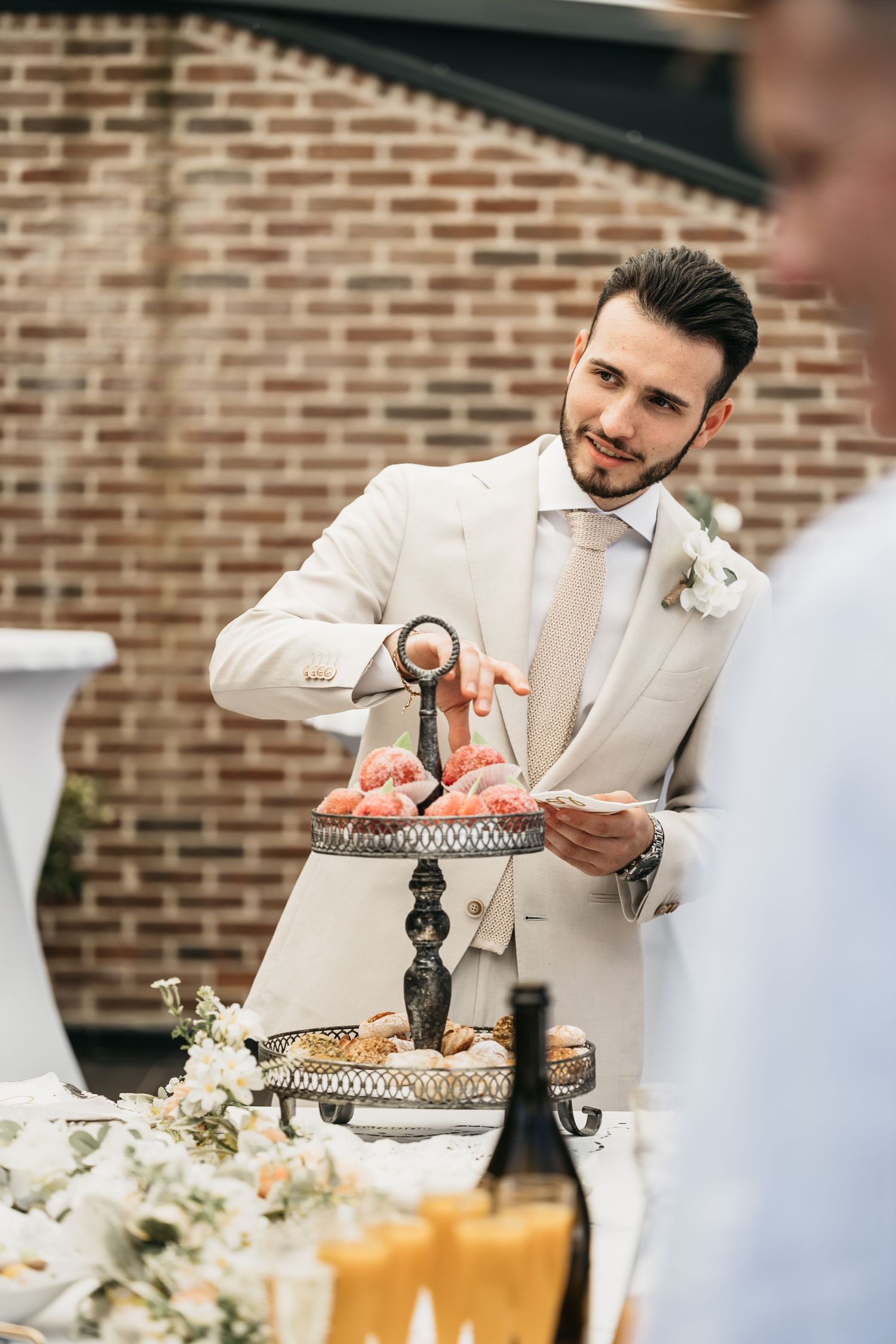 Dapper groom in beige suit selecting appetizers from decorative tiered tray at wedding reception.