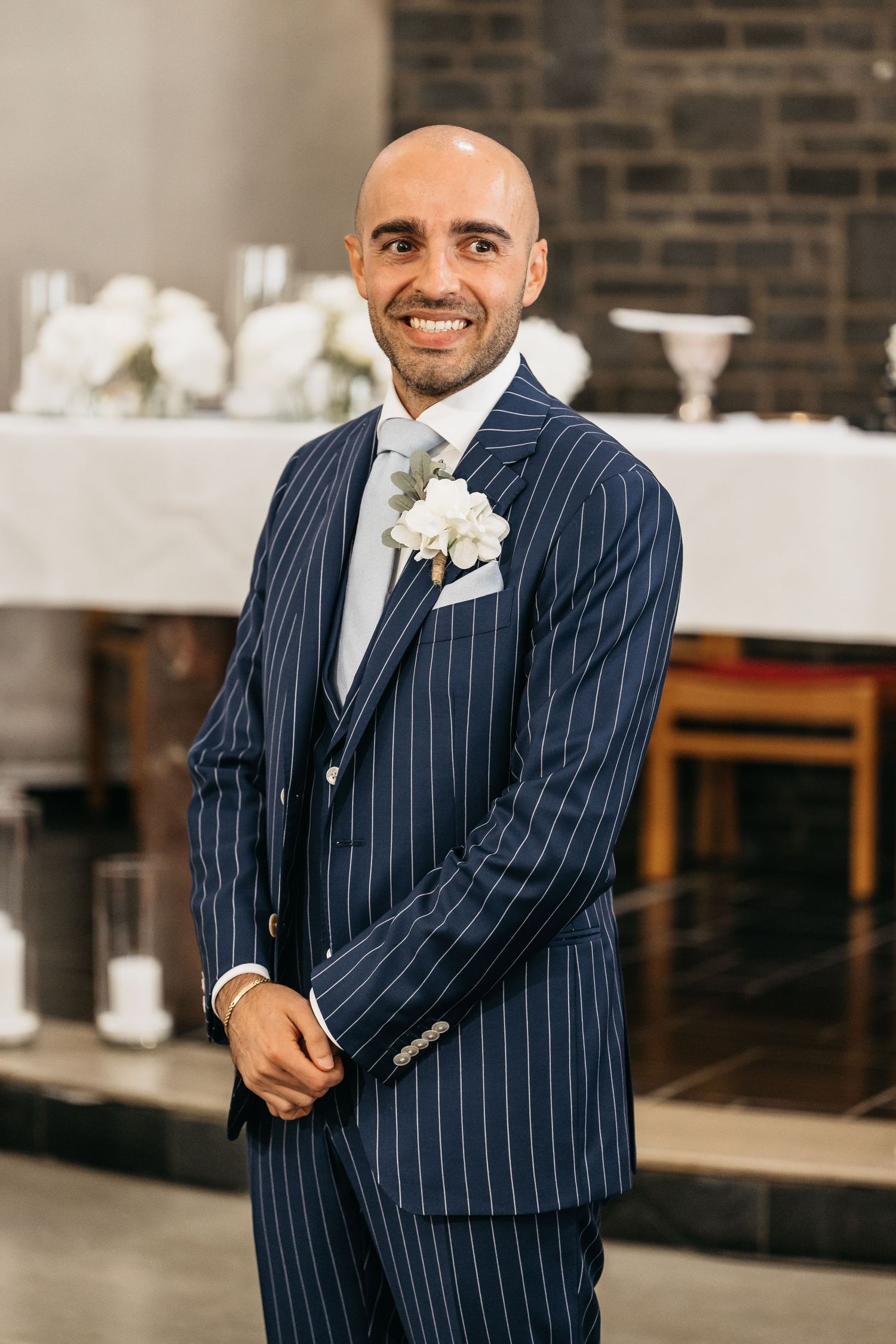 Smiling bald groom in a navy blue pinstripe suit with white boutonniere standing at church altar.