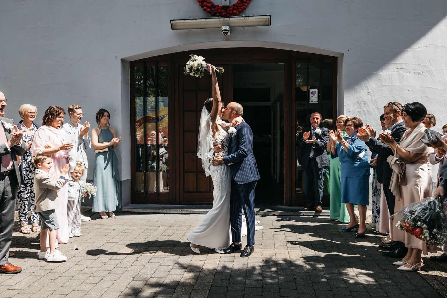 Bride and groom share romantic kiss outside wedding venue while guests celebrate around them.