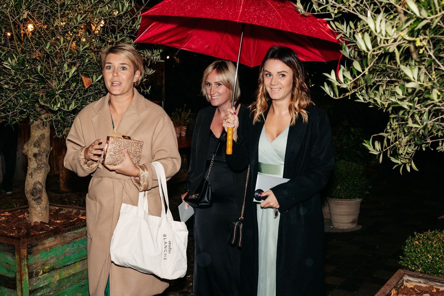 Three women standing outdoors at a wedding, one holding a red umbrella in the evening.