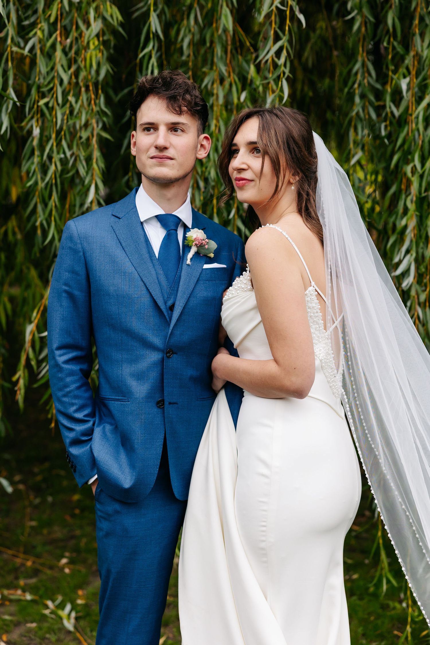 Bride in elegant white gown with veil poses with groom in blue three-piece suit under willow tree.