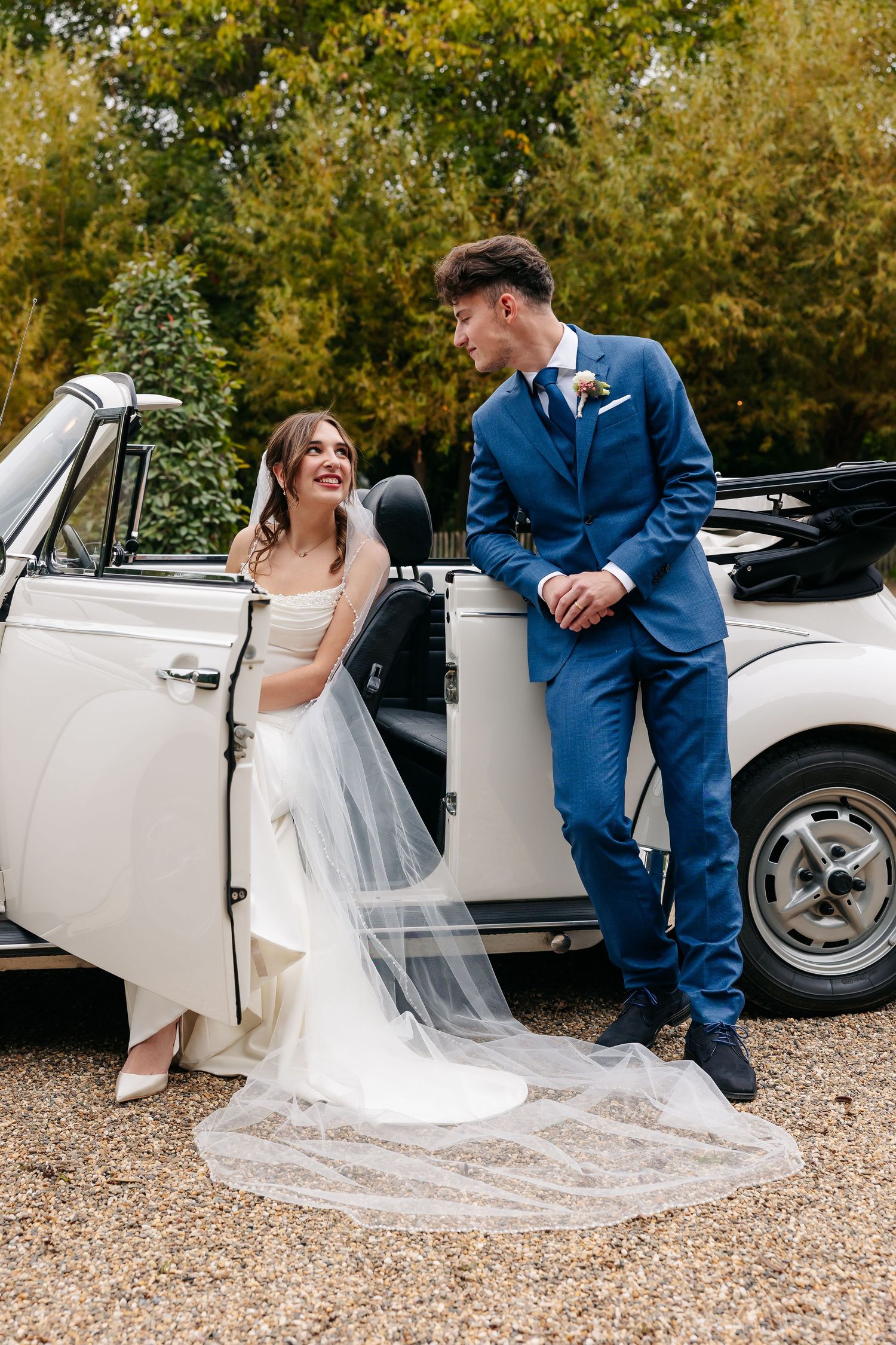 Bride in white gown with long veil and groom in blue suit smiling at each other by a vintage white car.