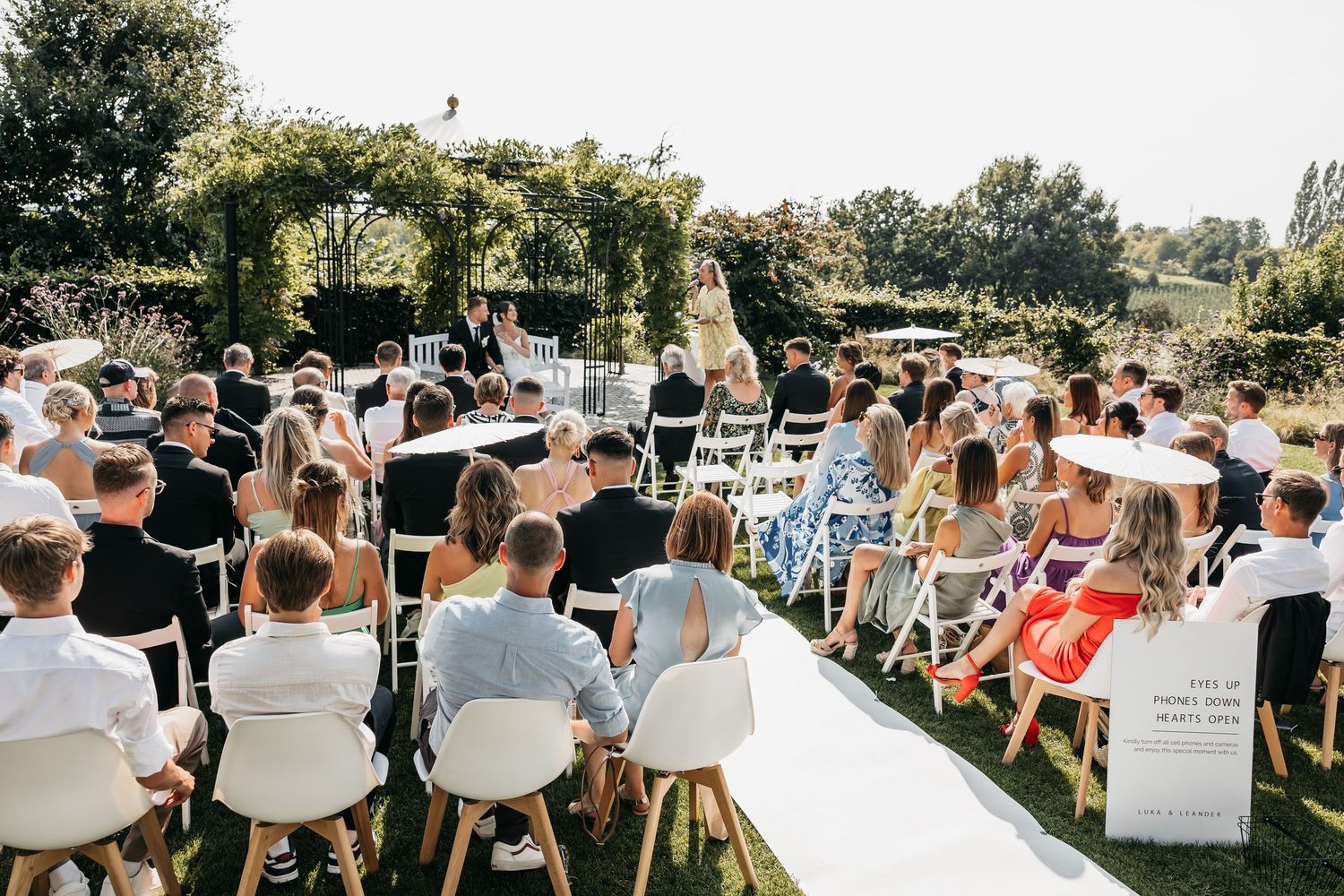 Outdoor wedding ceremony with guests seated on white chairs along a white aisle runner in a lush garden.