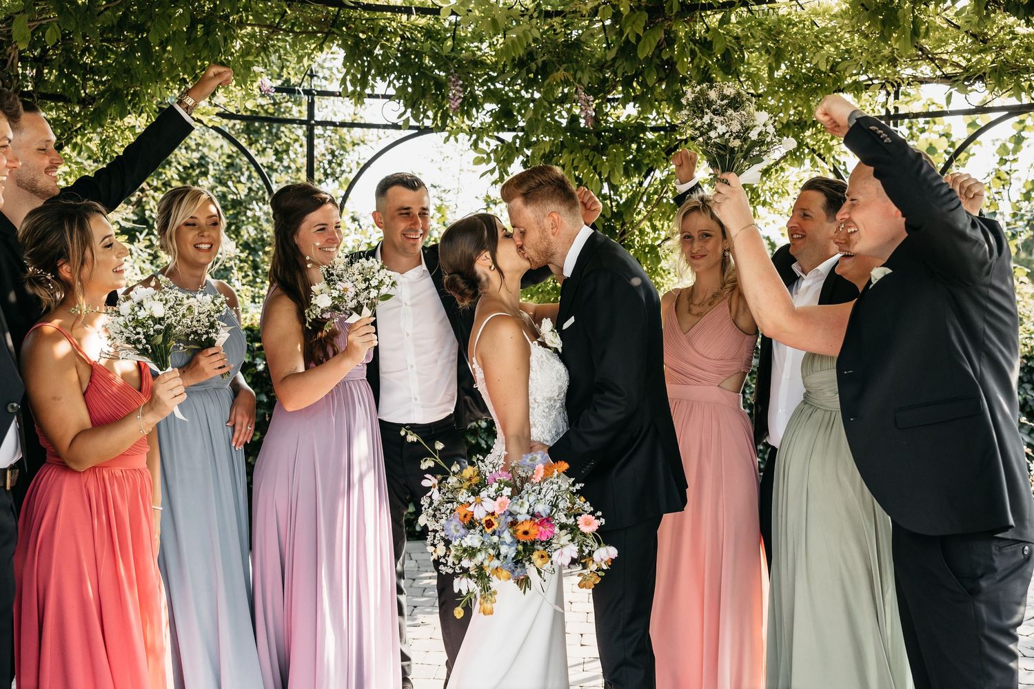 Wedding party celebrating with bride and groom kissing under a floral arch surrounded by bridesmaids and groomsmen.