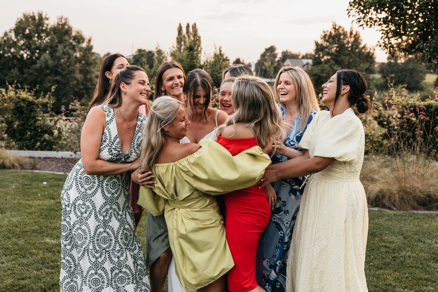 Group of women in colorful dresses laughing and hugging outdoors at a wedding celebration.