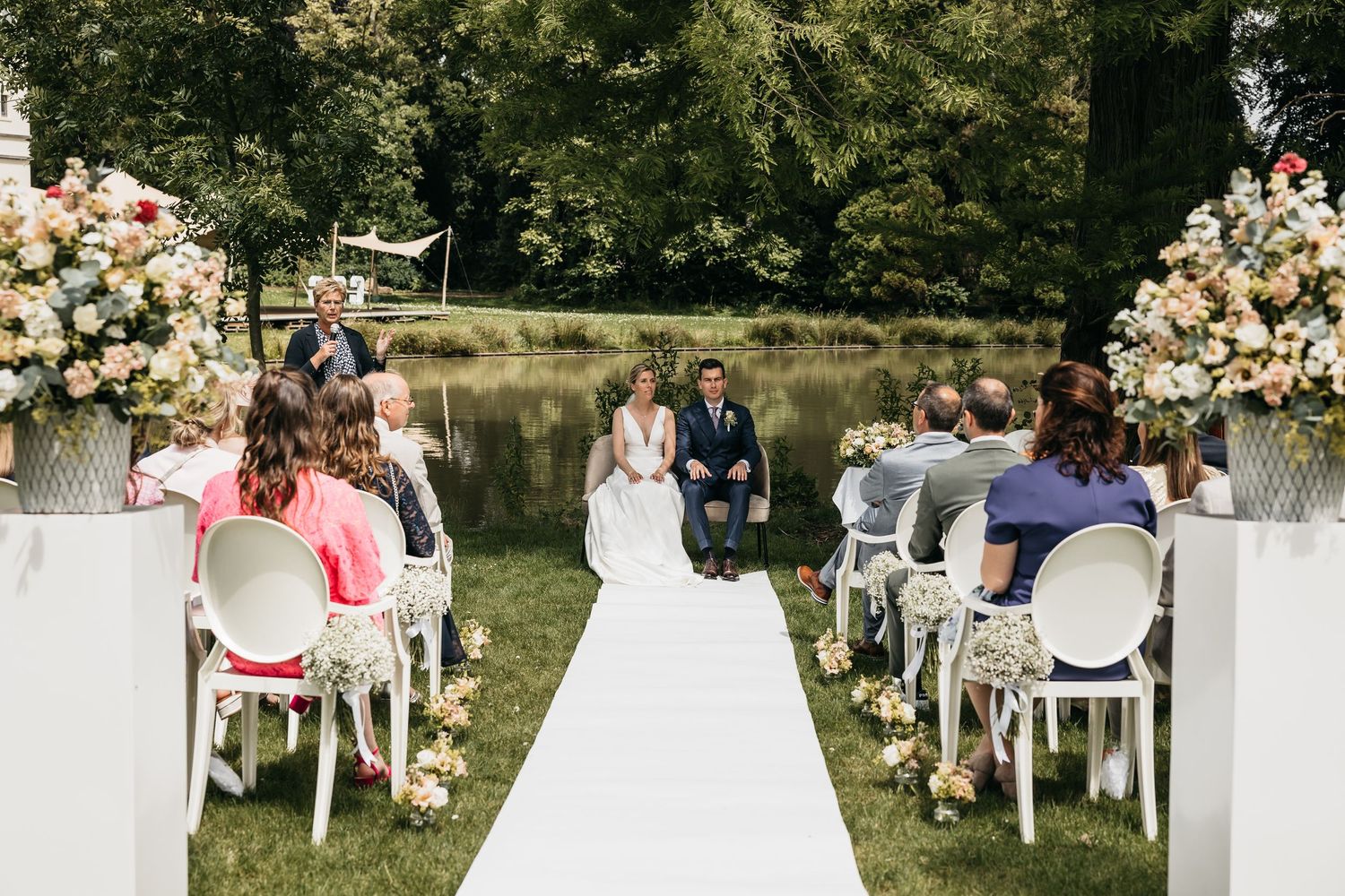 Elegant outdoor wedding ceremony with white aisle, floral arrangements, and guests seated by a pond.