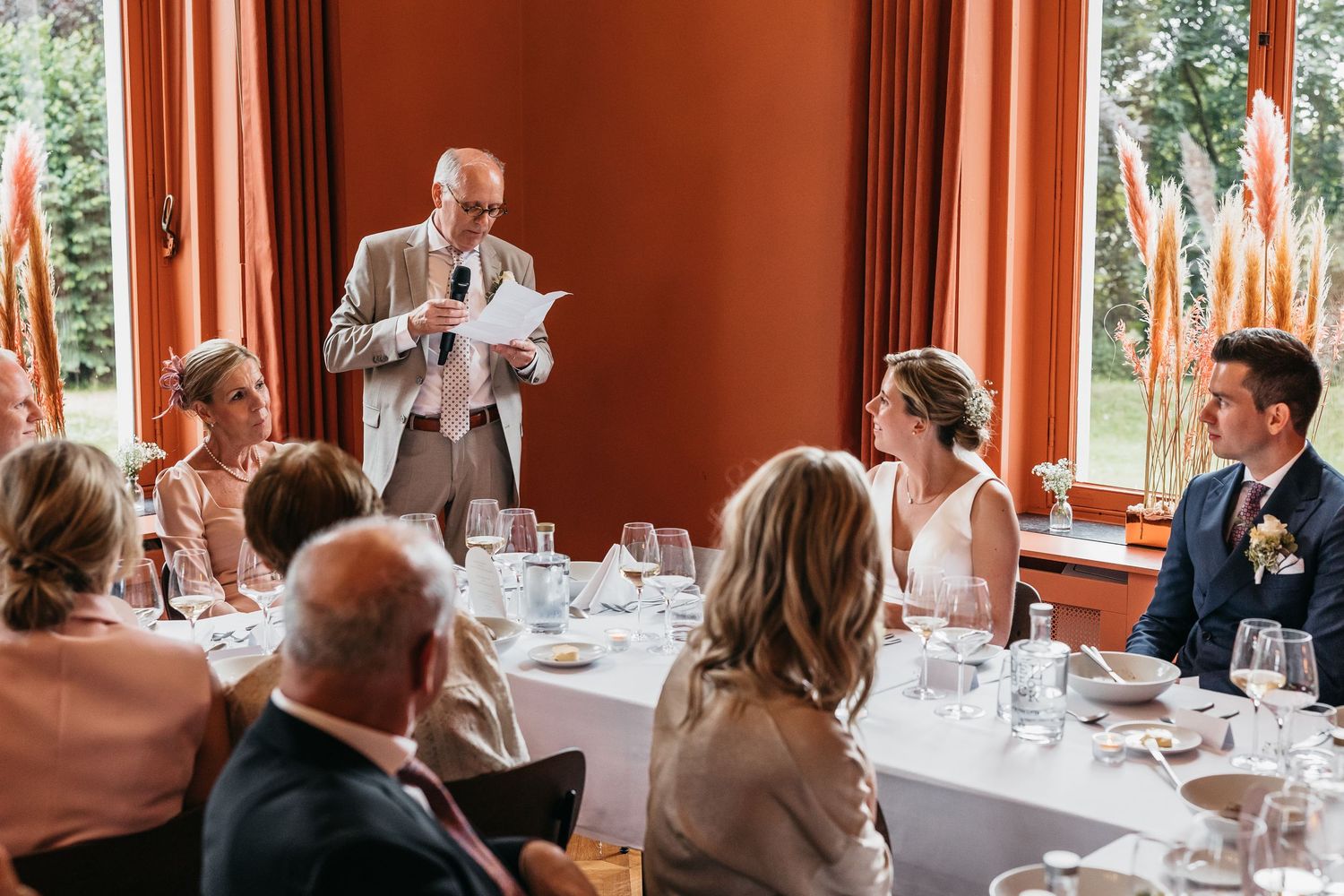 Older gentleman reads wedding speech to smiling bride and groom at reception dinner.