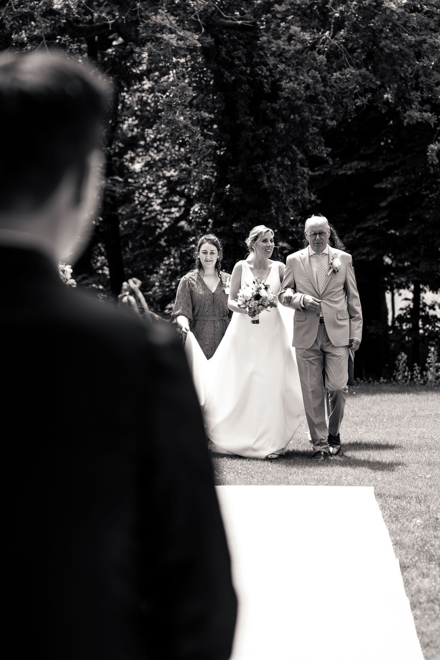 Black and white photo of bride walking down aisle with father and bridesmaid at outdoor wedding.