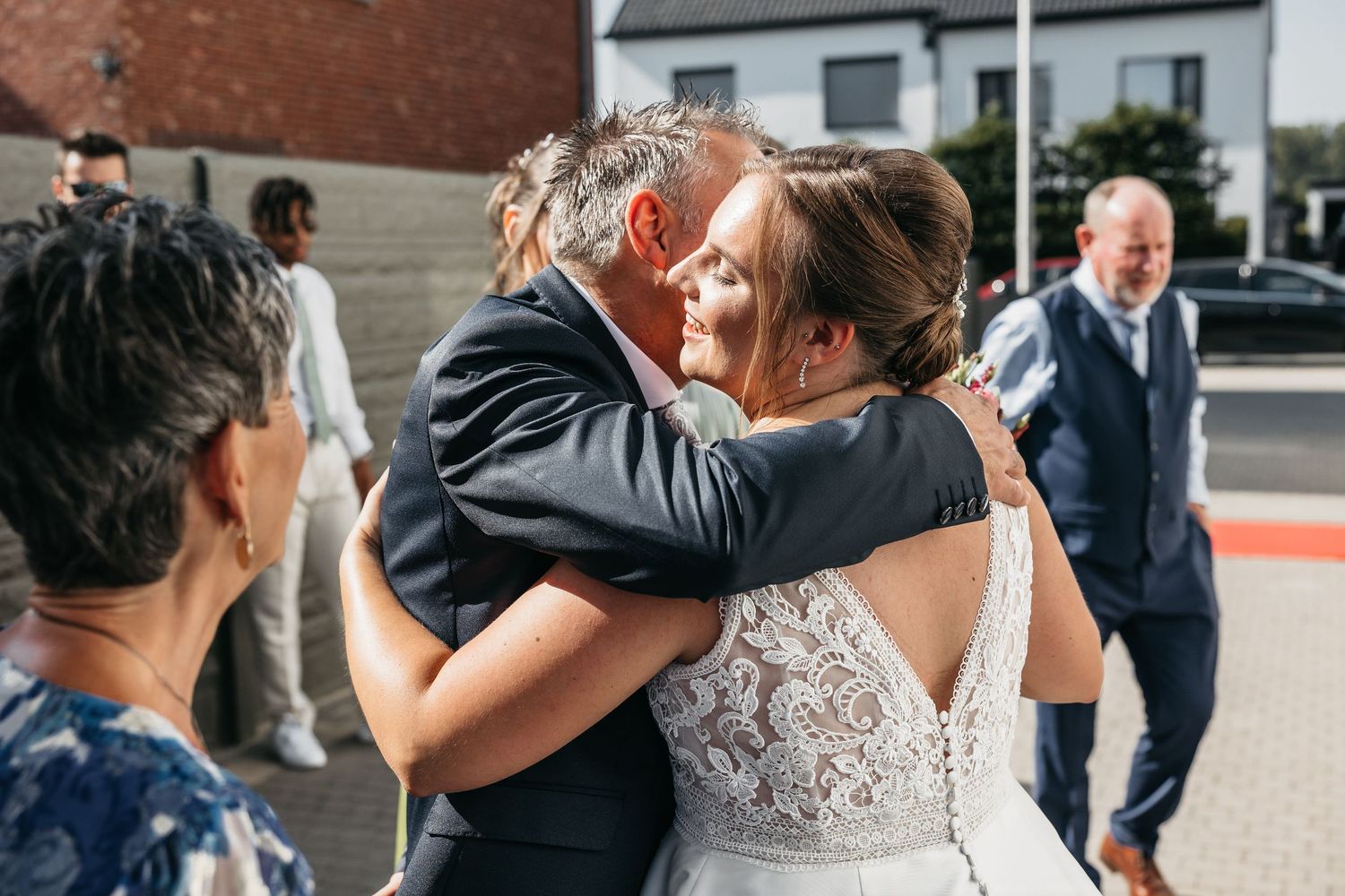 Bride in lace wedding dress shares a warm emotional hug with an older man at her wedding celebration.