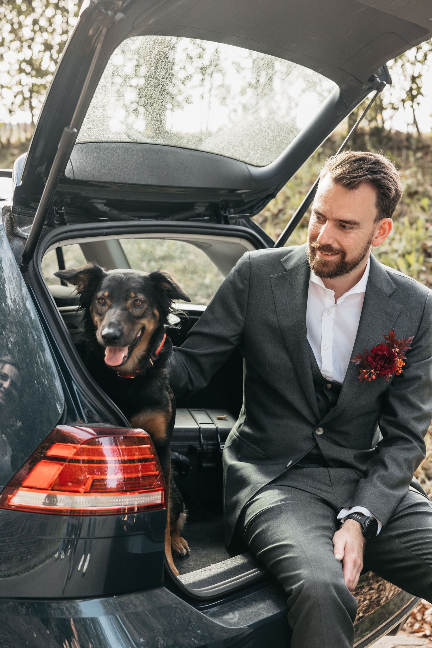 Groom in gray suit sitting on car trunk with happy black dog at outdoor wedding.