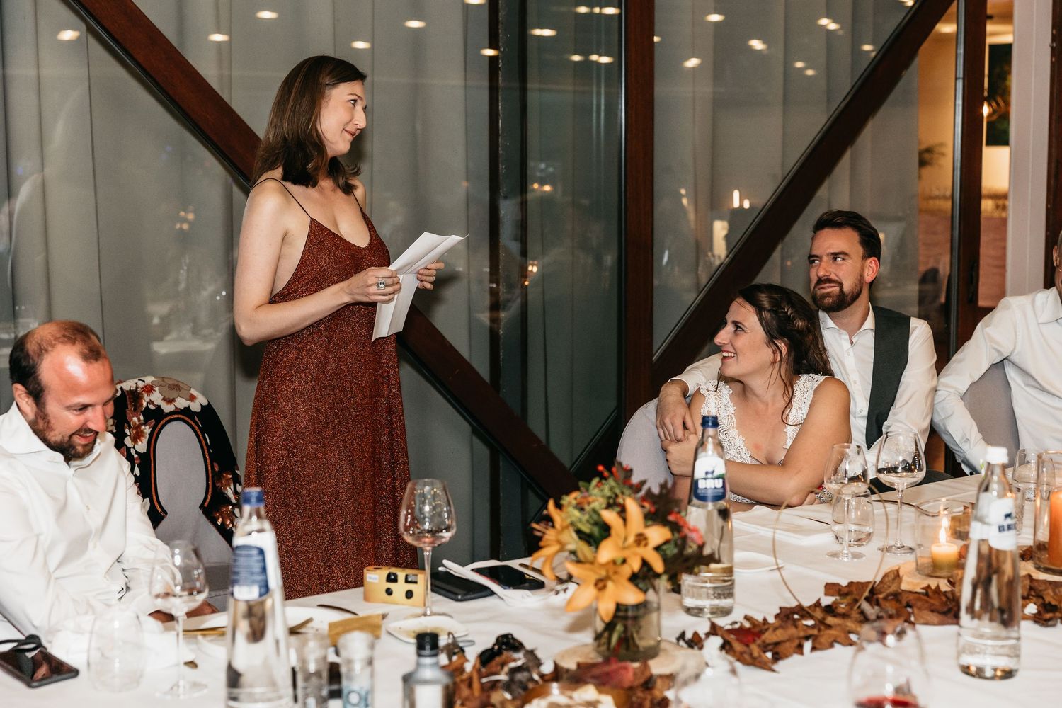 Woman in red dress giving a speech at a wedding reception while bride and groom listen.