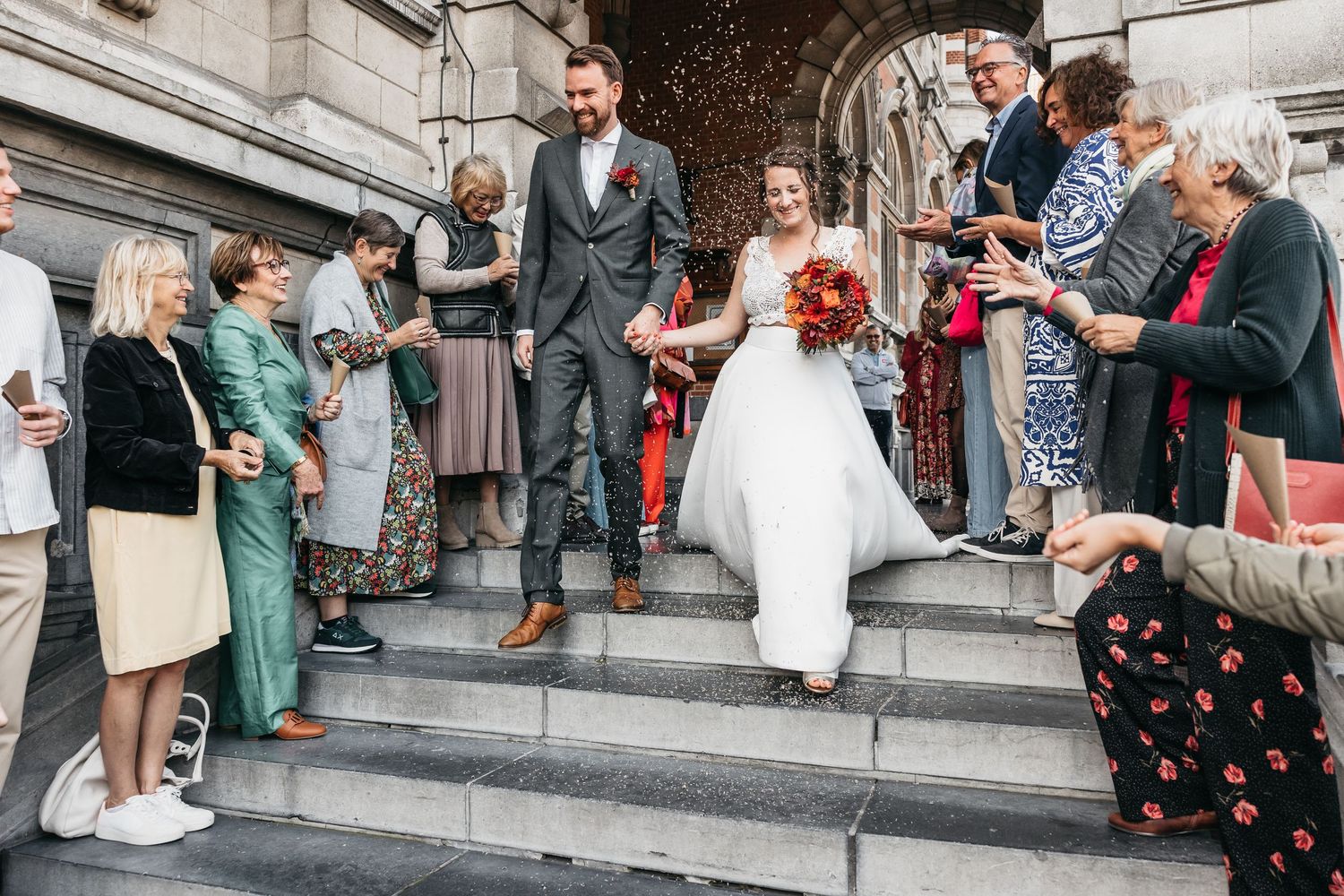 Joyful newlyweds descend stone steps as guests toss confetti in lively outdoor wedding celebration.
