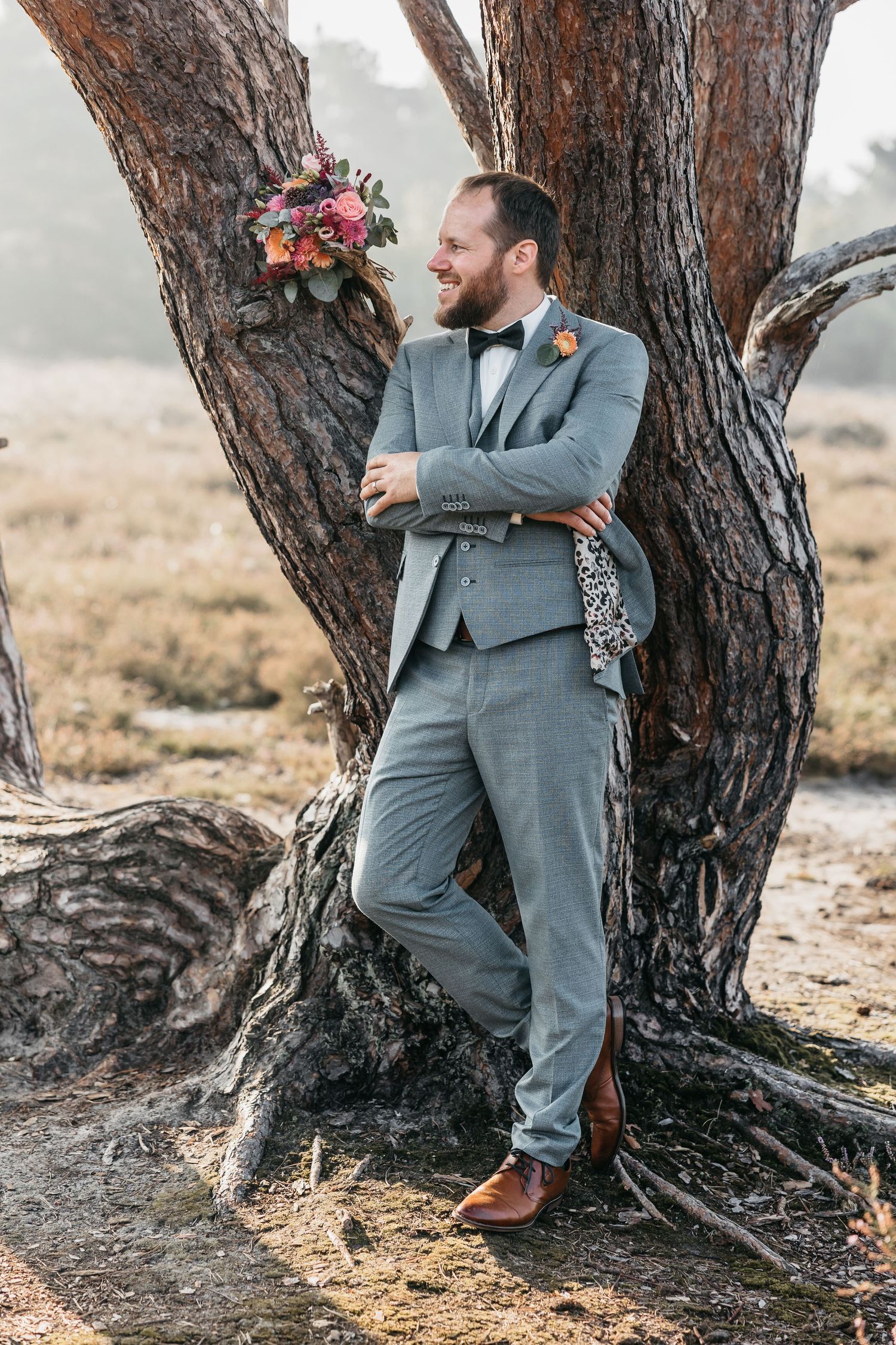 Groom in grey three-piece suit with bow tie leans against a pine tree with colorful bridal bouquet nearby.