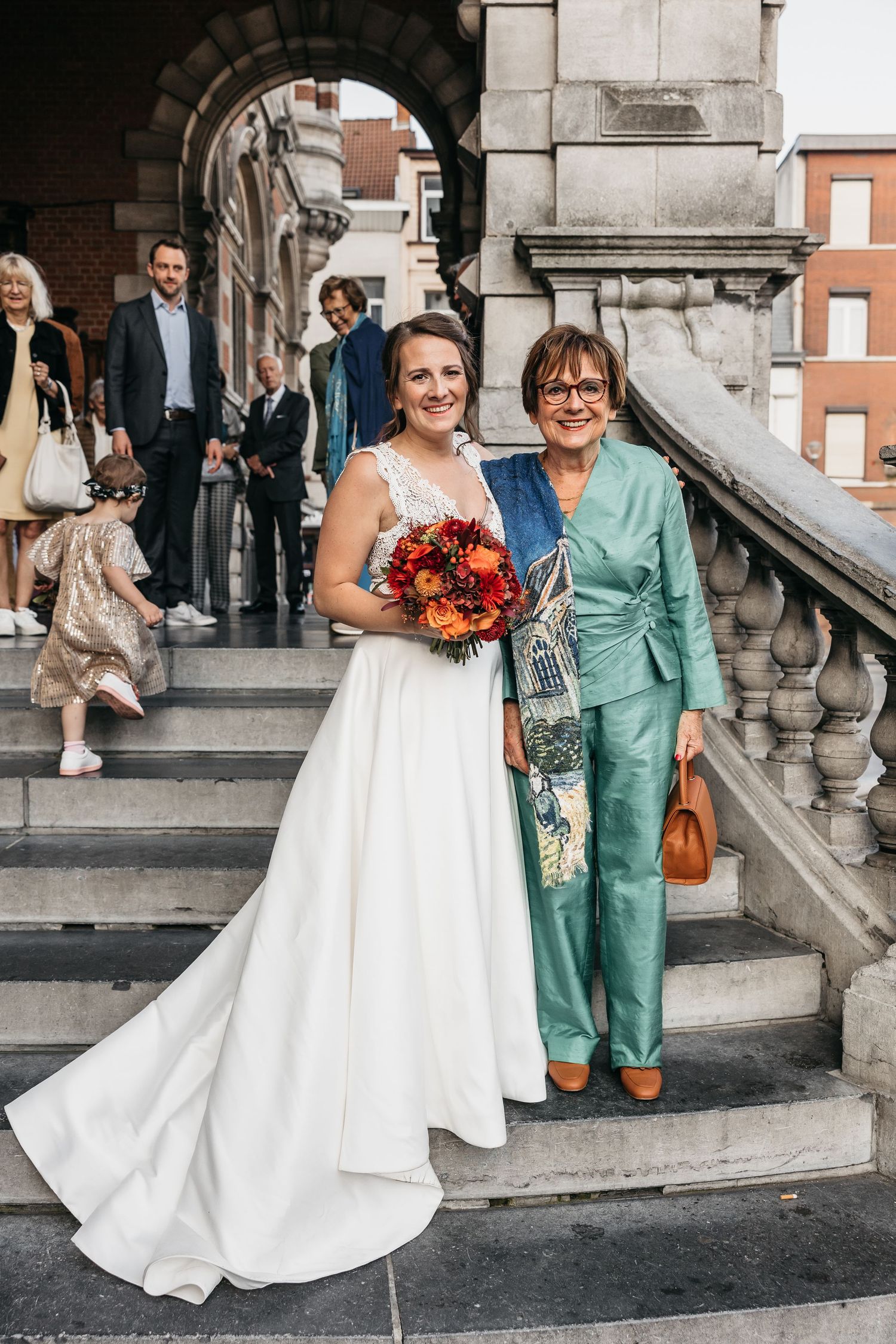 Bride in white lace gown holding autumn bouquet posing with older woman in teal suit on steps.