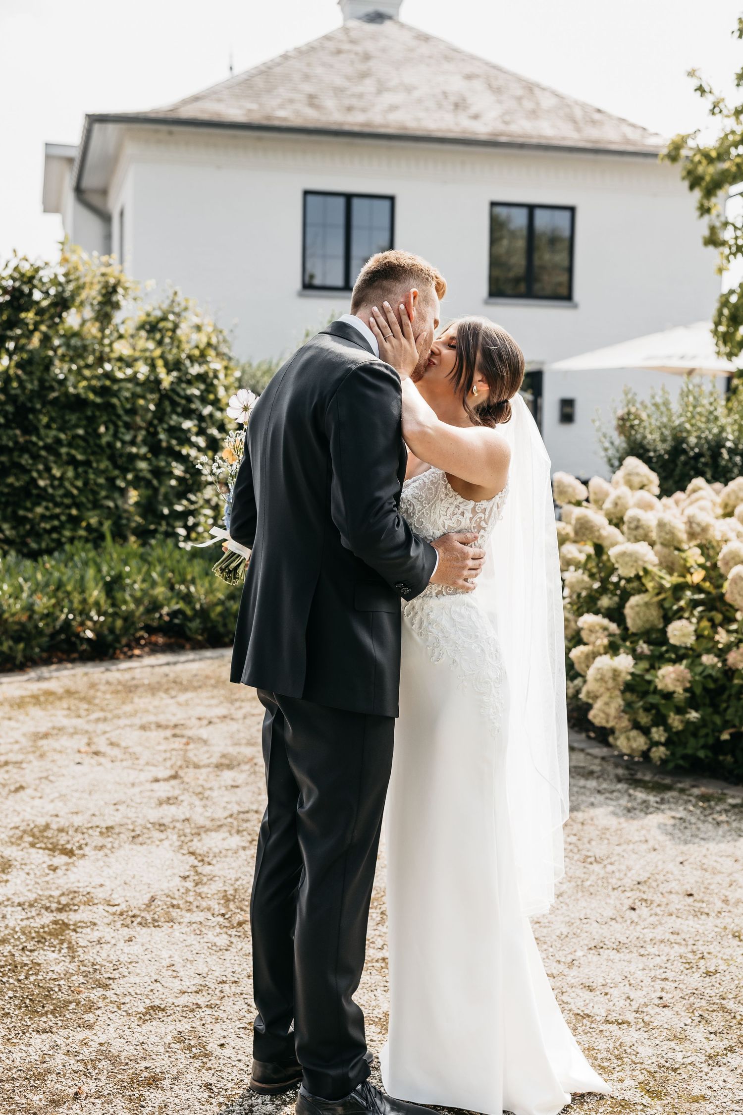 Bride and groom share romantic kiss in front of white country house with hydrangeas.