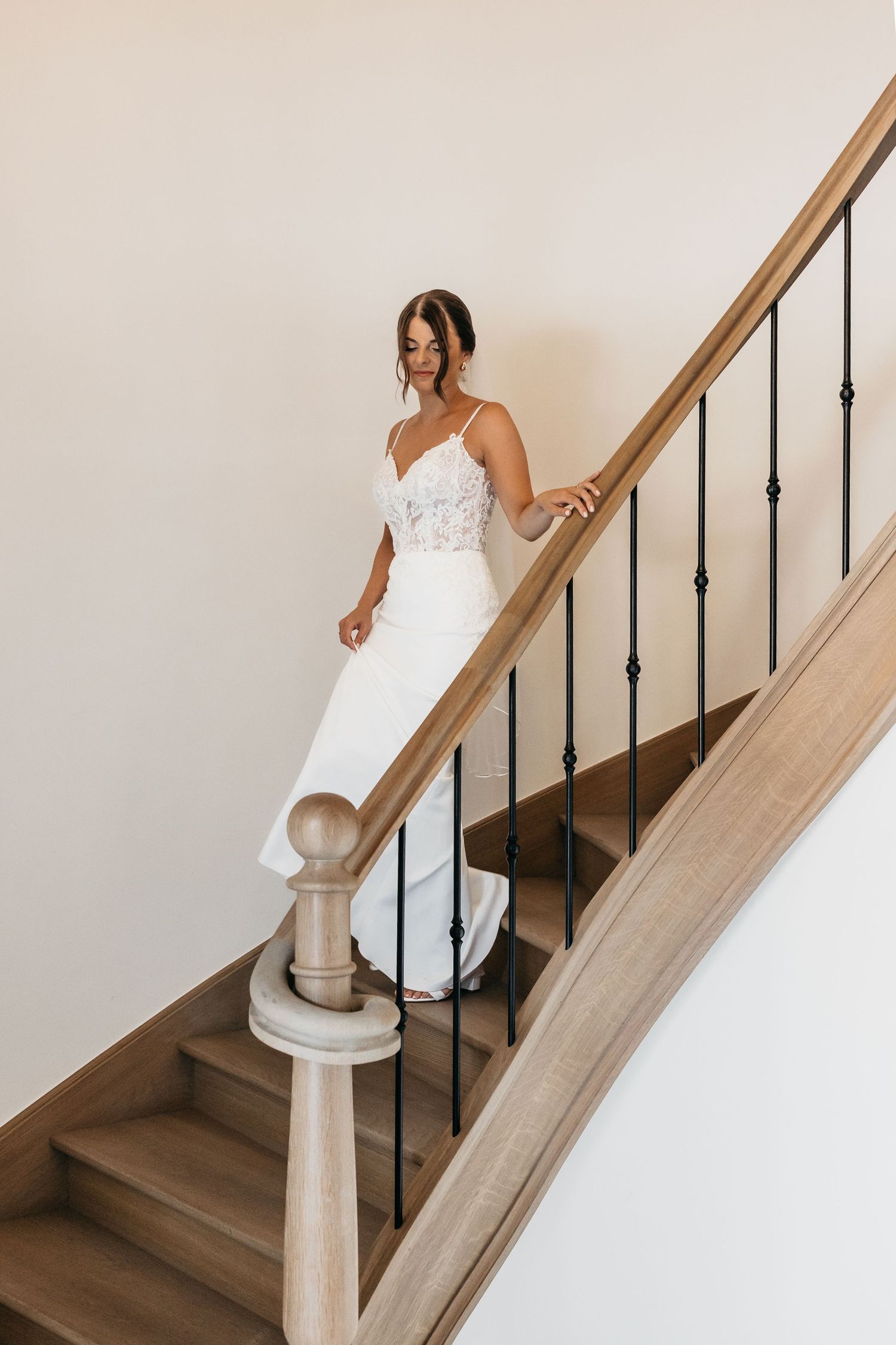 Bride in white lace wedding gown descending elegant wooden staircase with black iron balusters.
