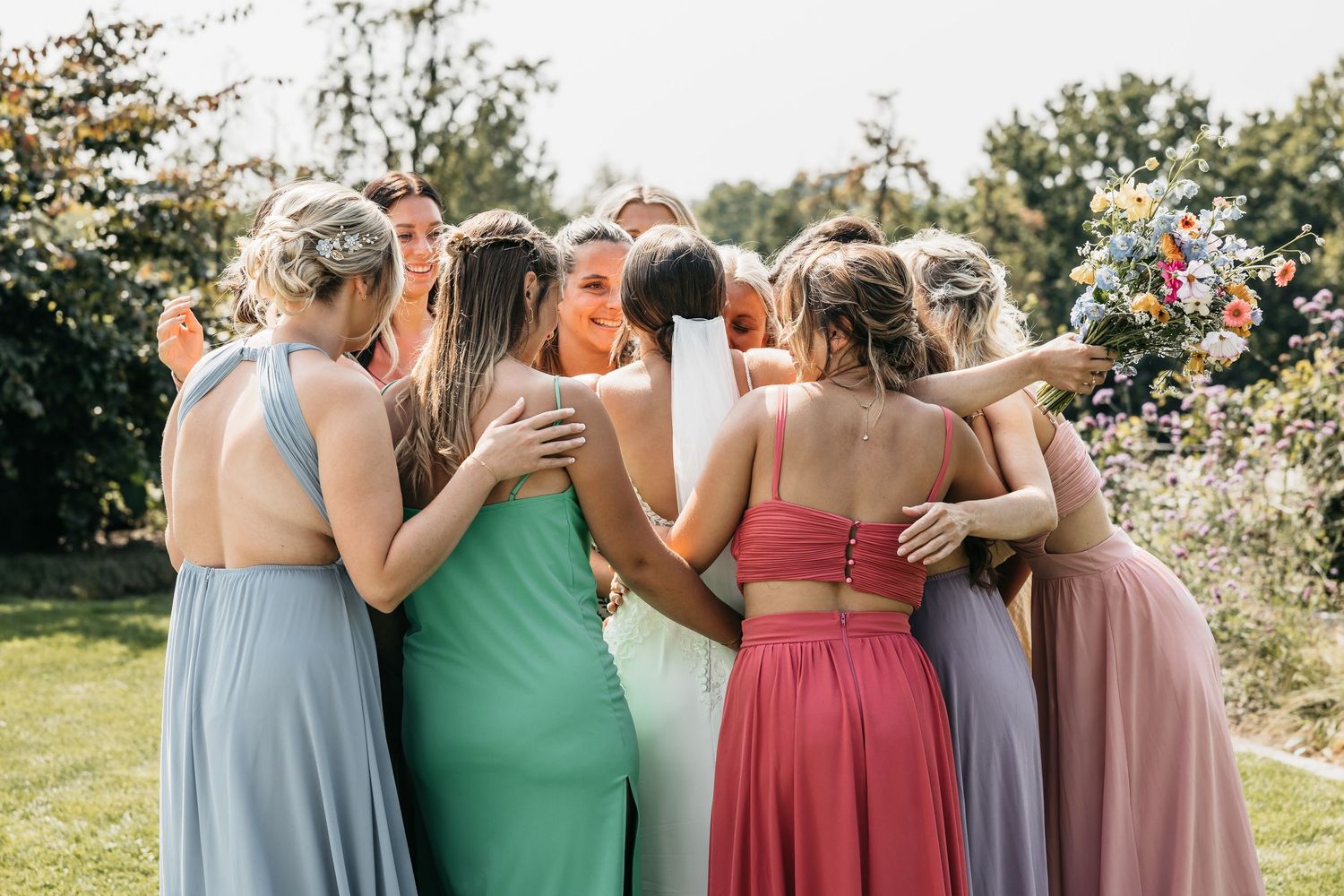 Bridesmaids in pastel dresses share an emotional group hug with the bride in a sunny outdoor garden setting.