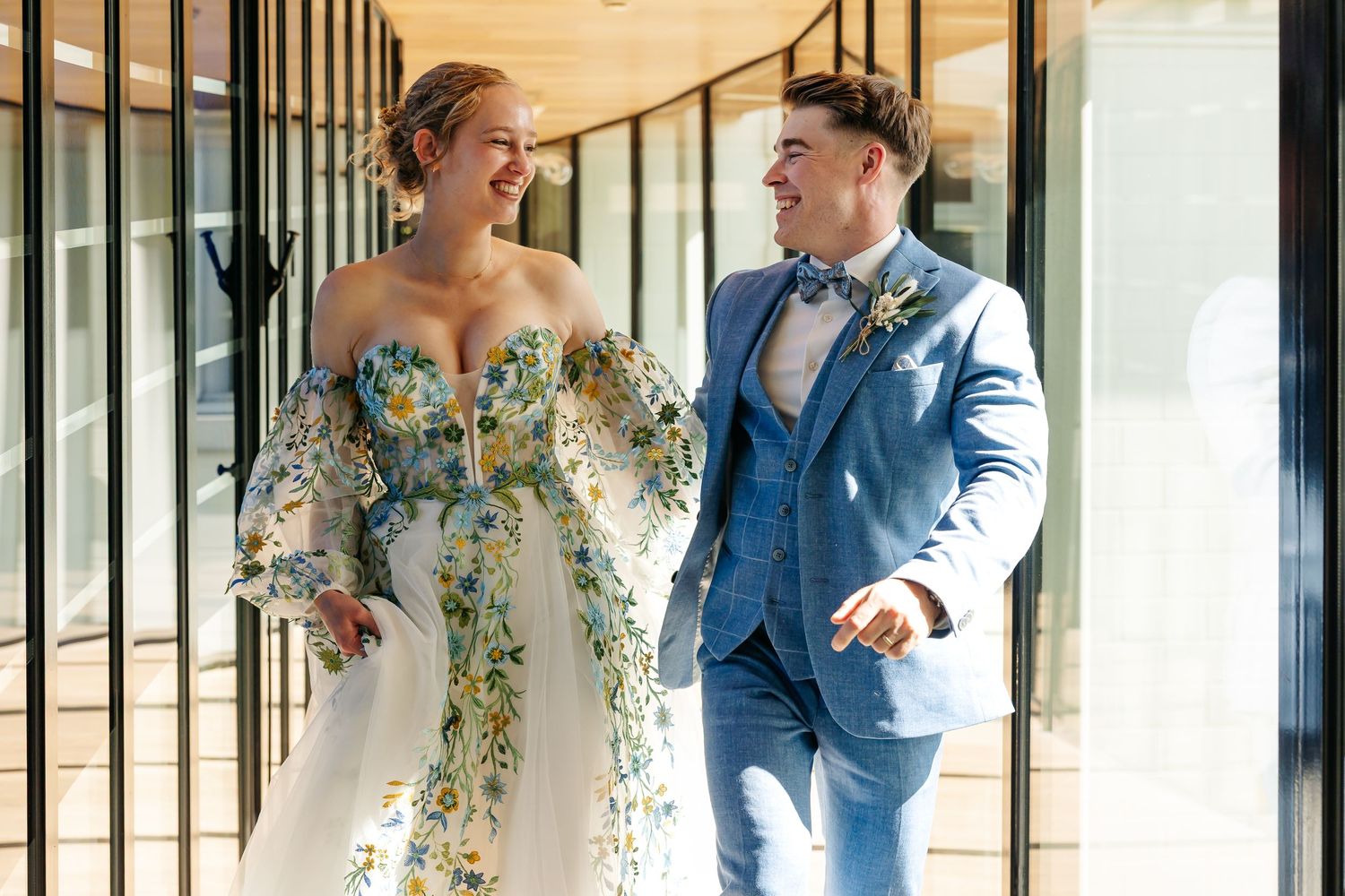 Laughing bride in off-shoulder floral gown and groom in light blue suit walking together at wedding.