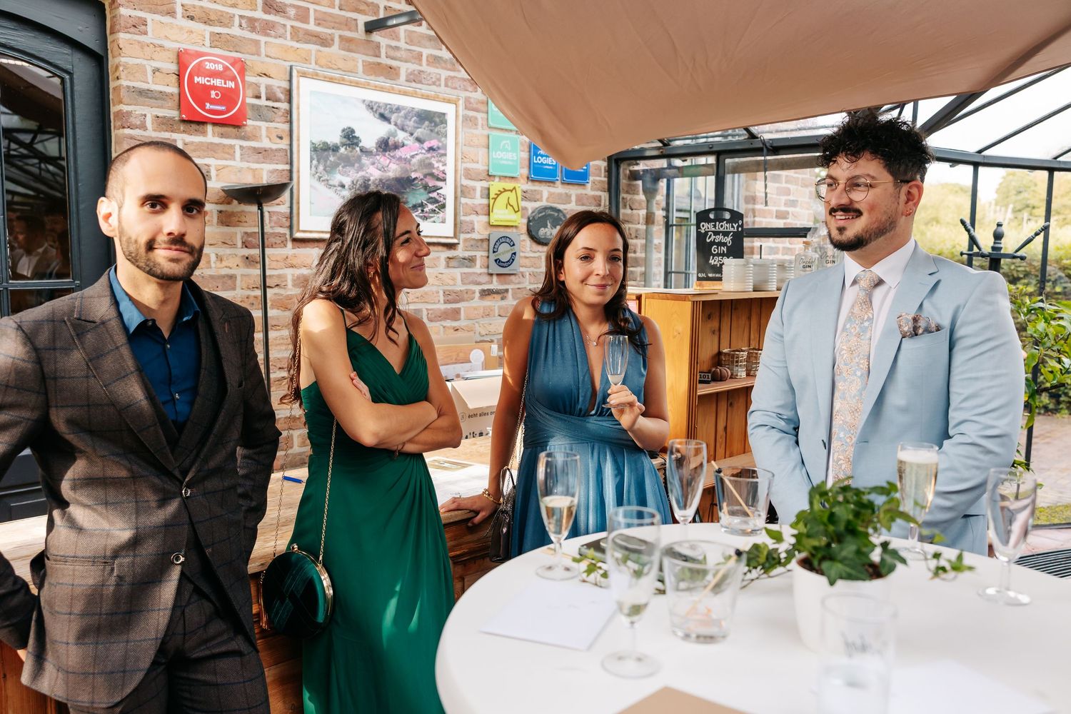 Four elegantly dressed wedding guests socializing around a white table in a rustic brick venue.
