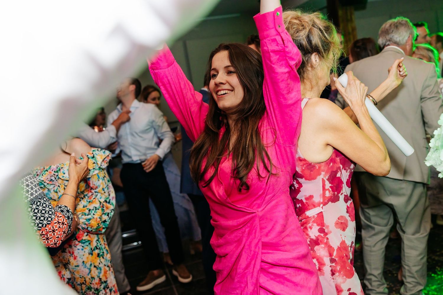 Woman in bright pink dress dances joyfully with arms raised at a lively wedding reception party.