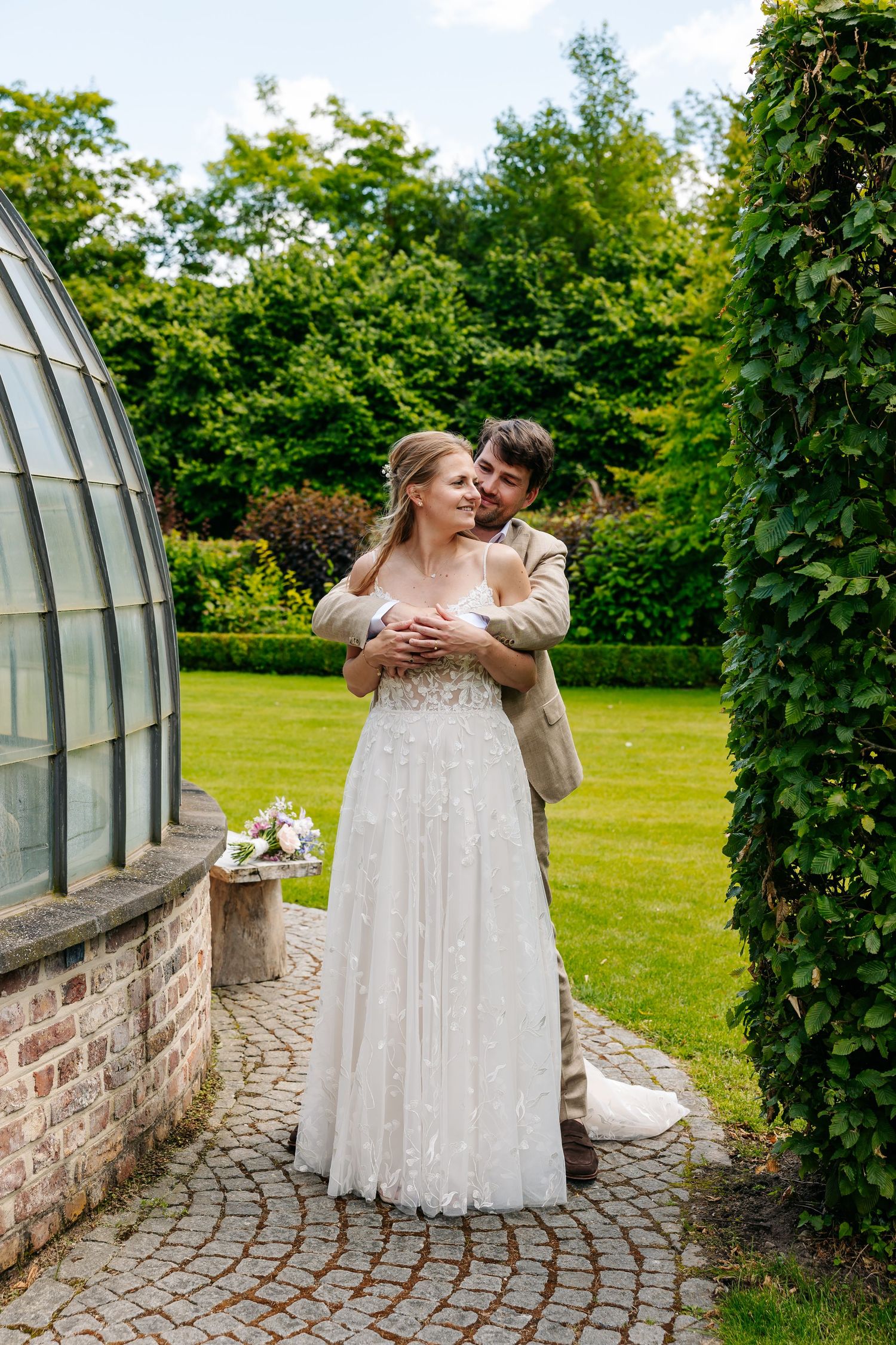 Groom in beige suit embracing bride in white lace gown in lush garden setting.