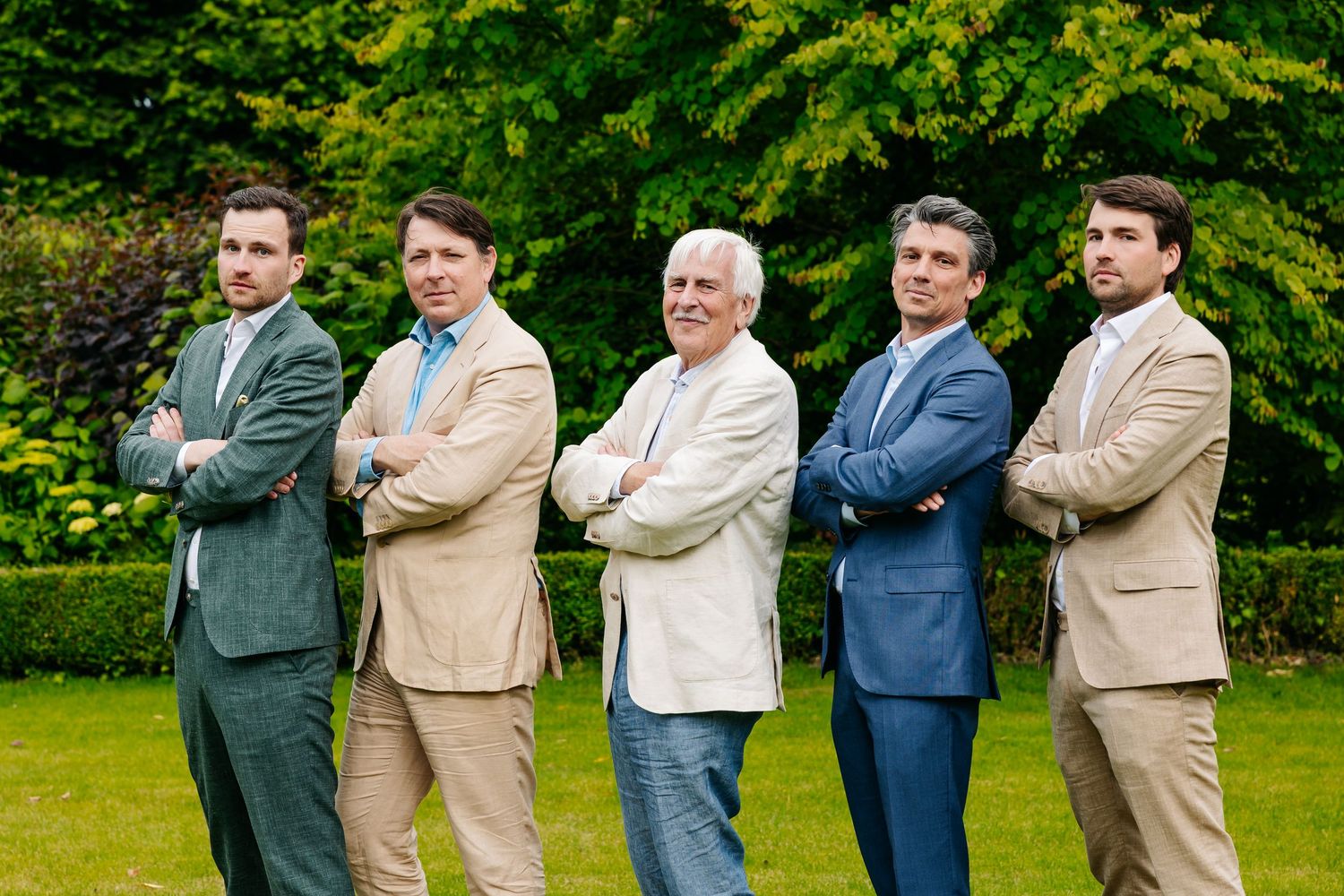 Five men in suits and blazers posing with crossed arms in a green garden at a wedding celebration.