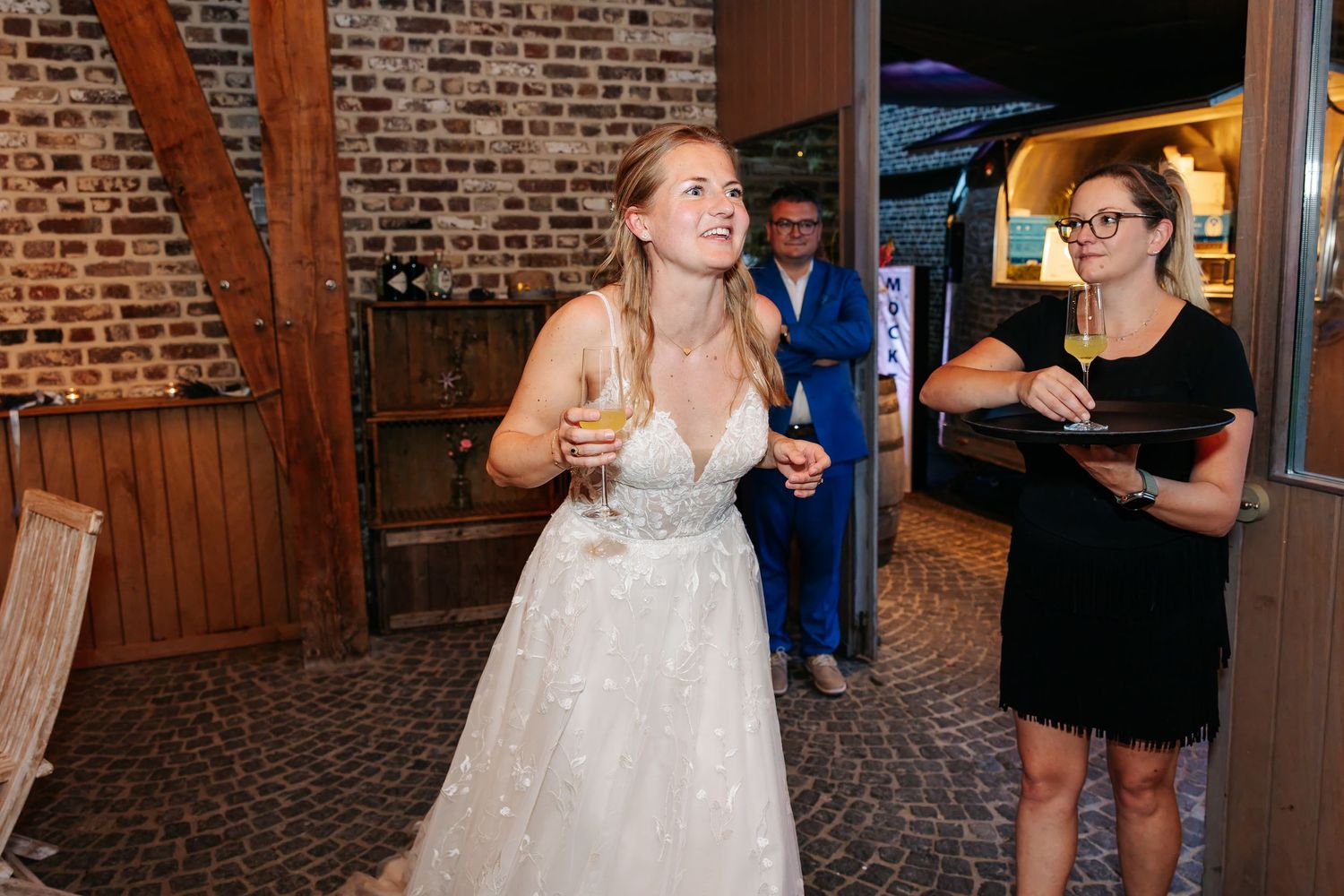 Bride in white wedding gown holds champagne glass while woman in black serves at elegant rustic venue.
