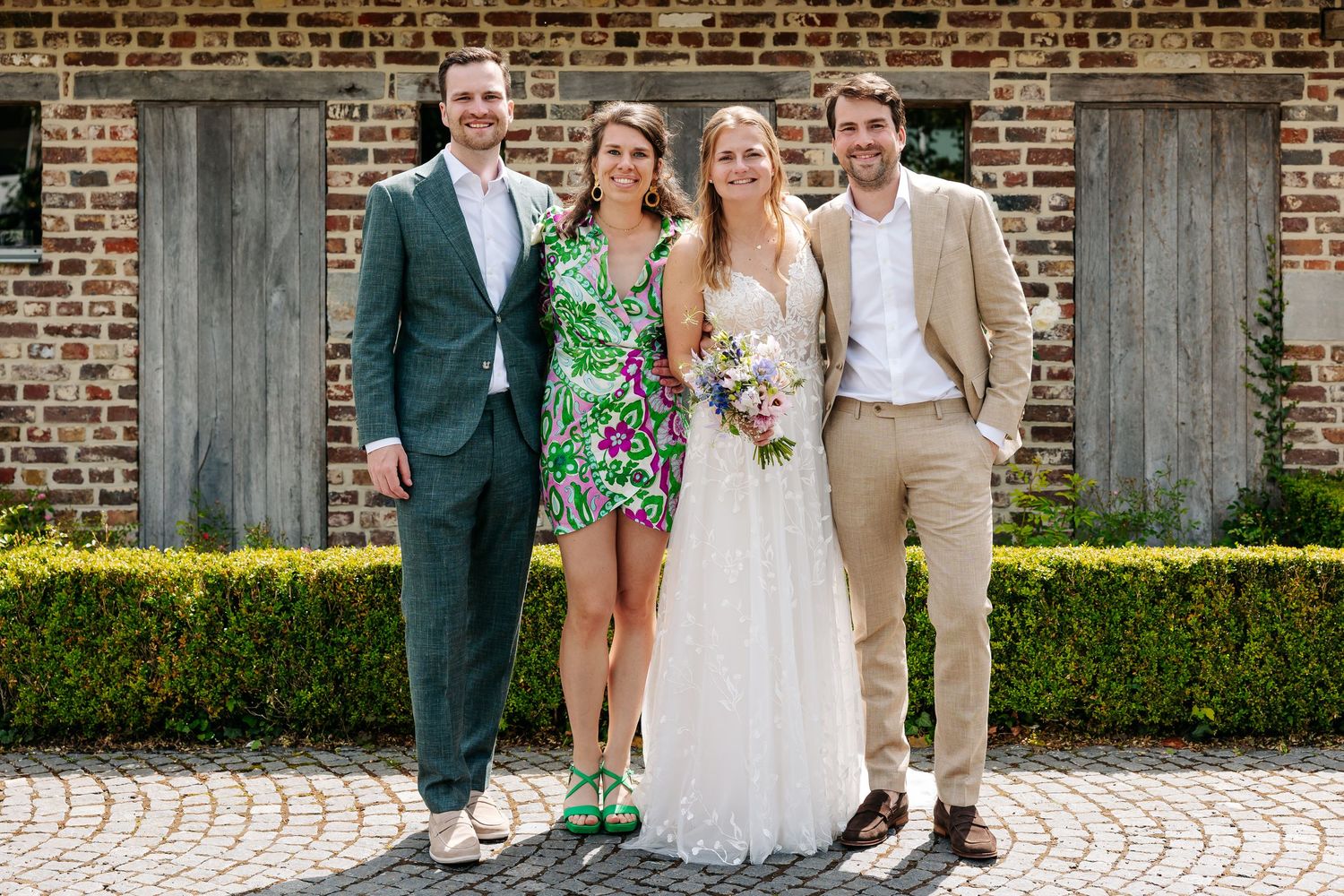 Bride in lace gown and groom in beige suit posing with two guests in front of a brick courtyard.