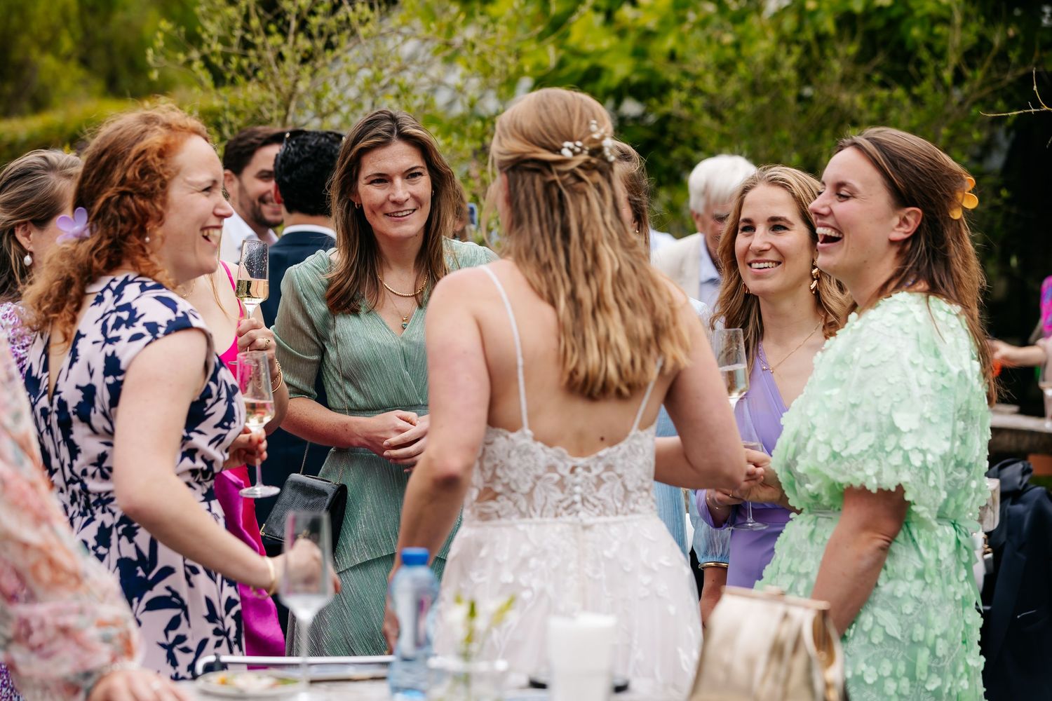 Bride in lace wedding dress laughing with female guests at outdoor wedding reception.