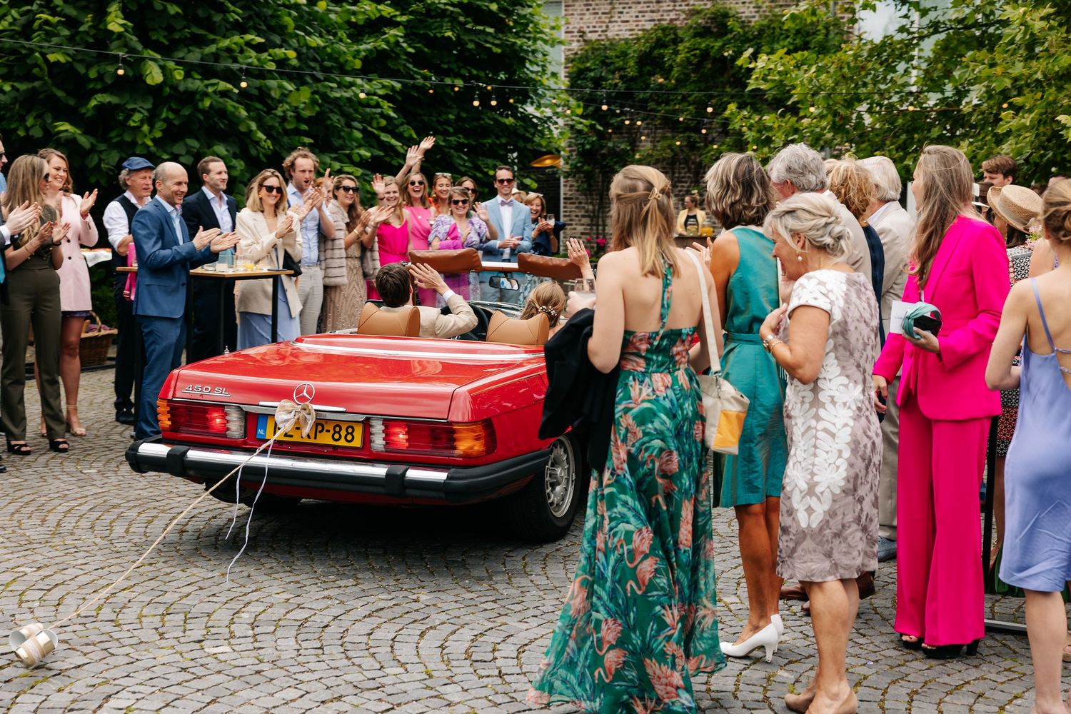 Wedding guests gather around a red vintage Mercedes convertible on a cobblestone driveway during an outdoor celebration.