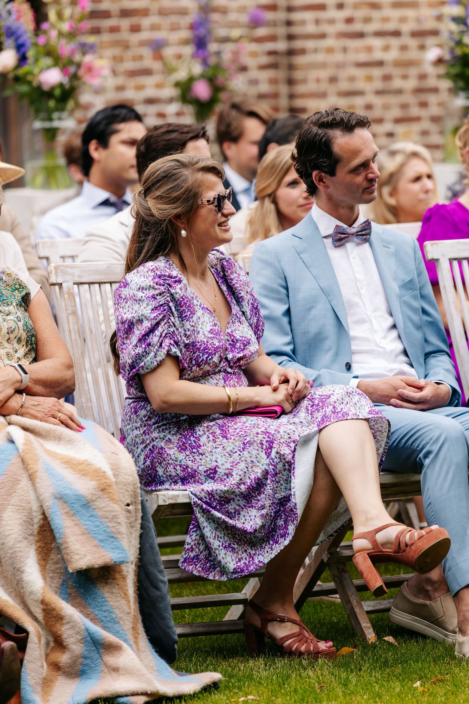 Wedding guests seated outdoors, woman in purple floral dress laughing beside man in light blue suit with bow tie.