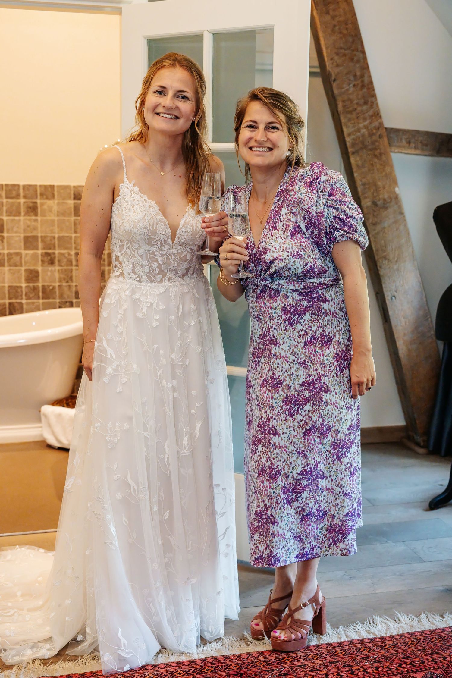 Smiling bride in white lace gown and woman in floral dress toasting with champagne.