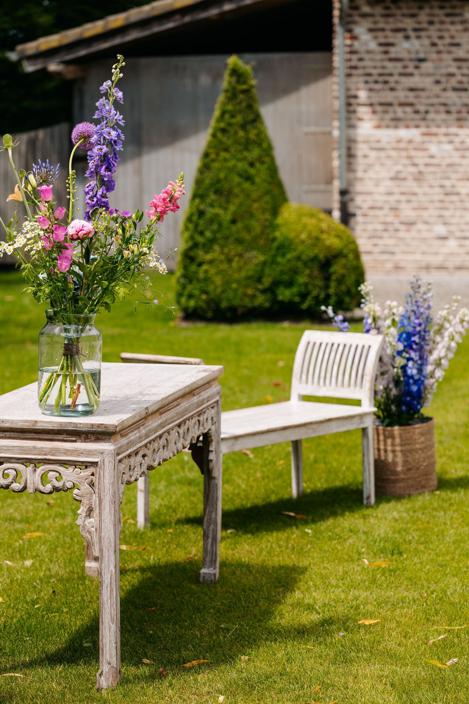 Elegant outdoor wedding setup with vintage white table, colorful wildflower bouquet, white bench, and manicured garden hedges.