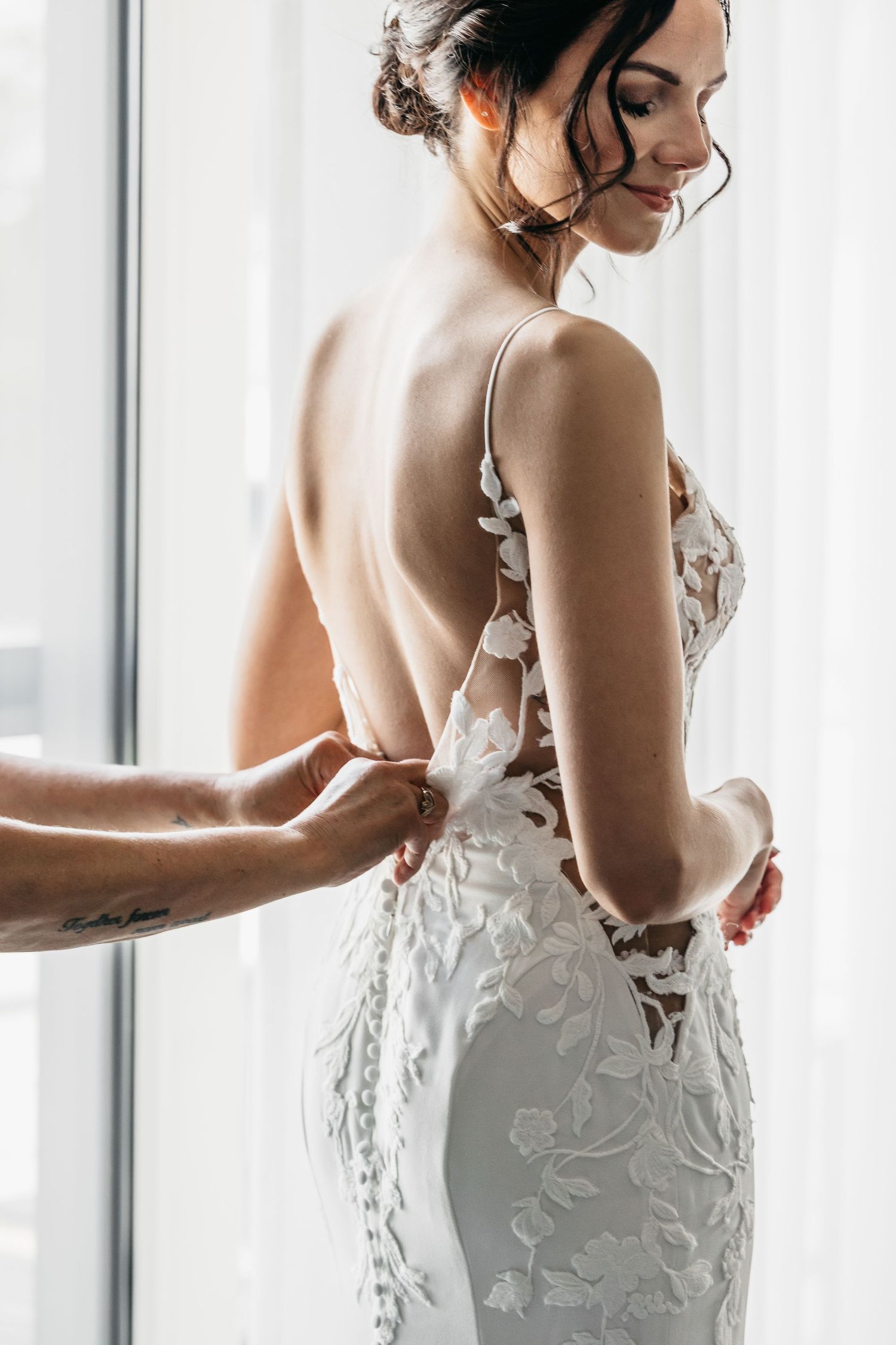 Bride in backless lace wedding gown being fastened by assistant near bright window.