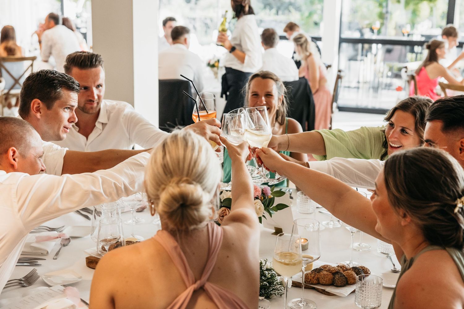 Smiling wedding guests clinking glasses in a joyful toast at a sunlit reception venue.