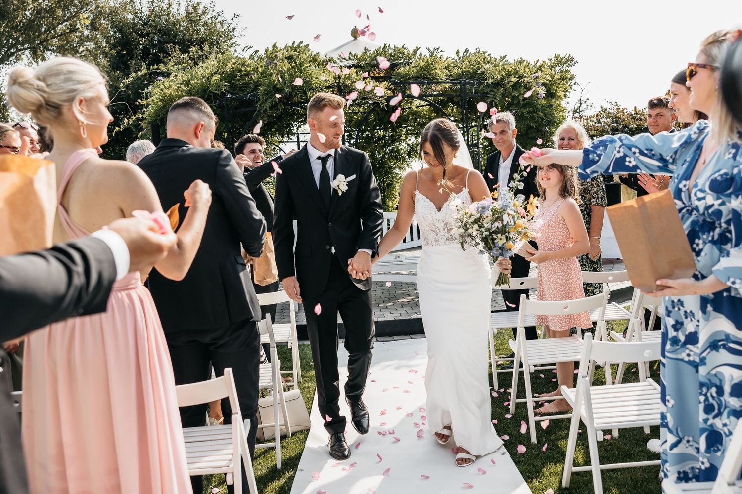 Bride and groom walk down the aisle as guests toss flower petals at outdoor wedding ceremony.
