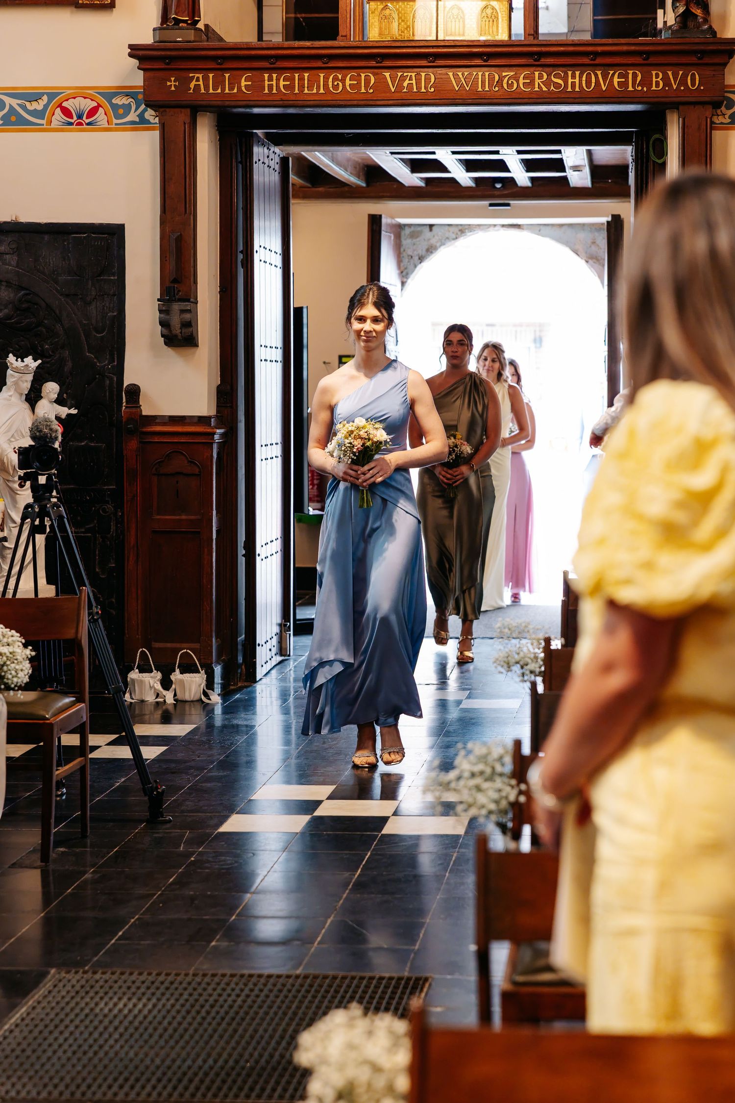 Bridesmaids in blue and green dresses walking down church aisle at wedding.