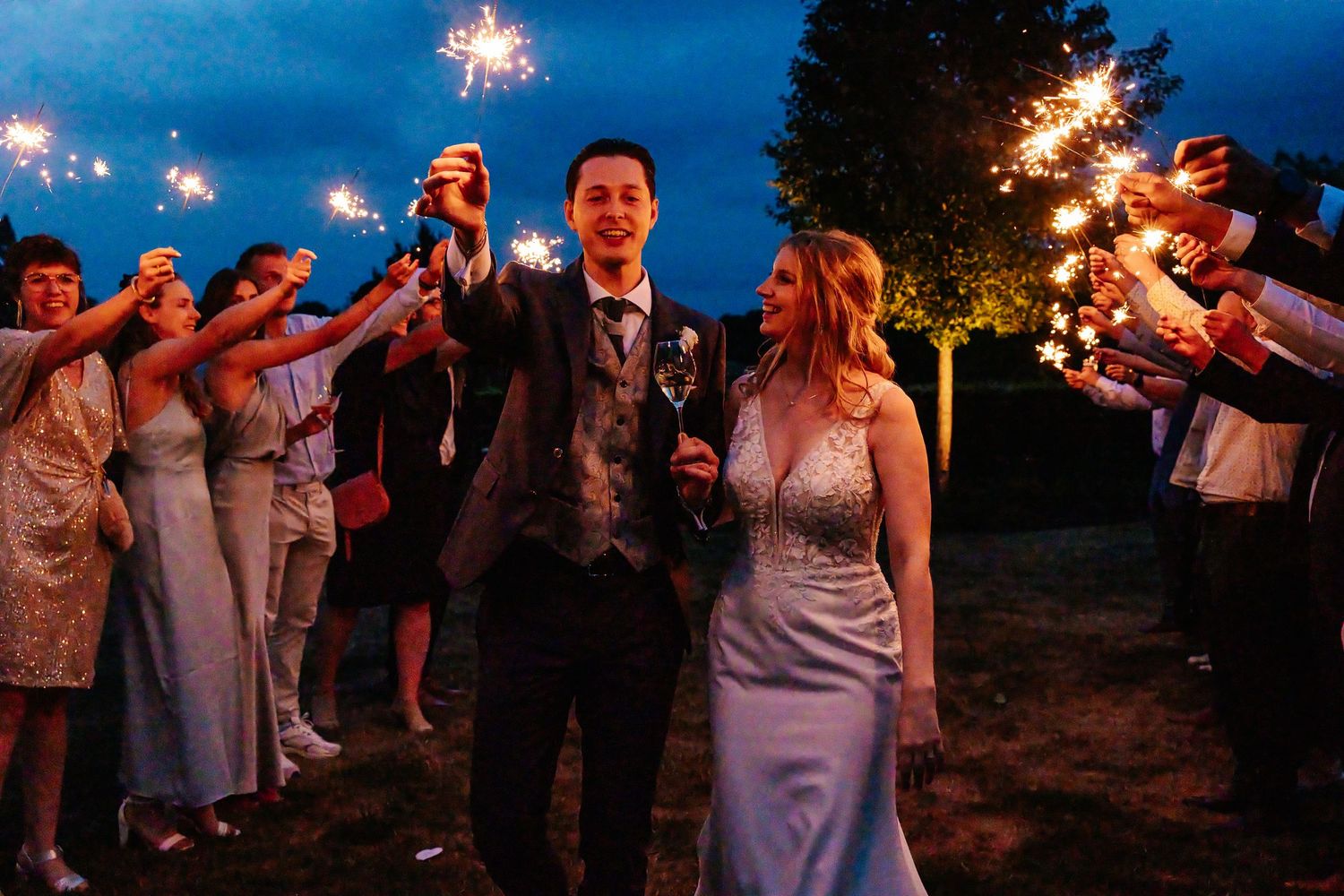Bride and groom walk through sparkler tunnel with wedding guests at twilight outdoor celebration.