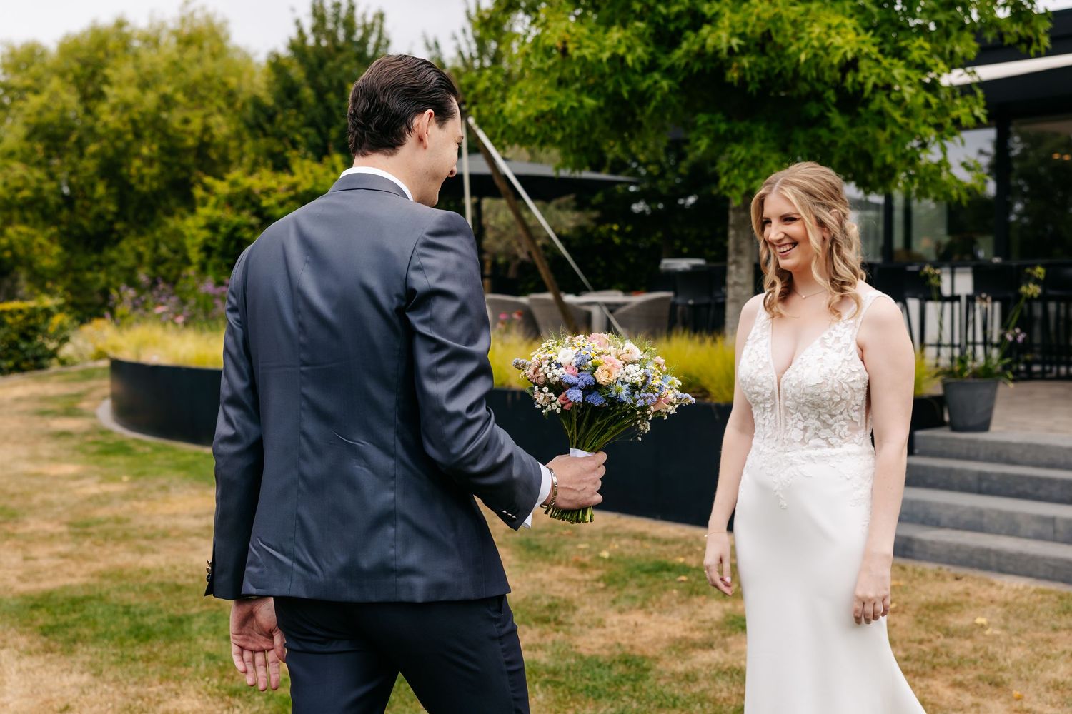 Groom turning to present colorful wedding bouquet to smiling bride in white gown outdoors.