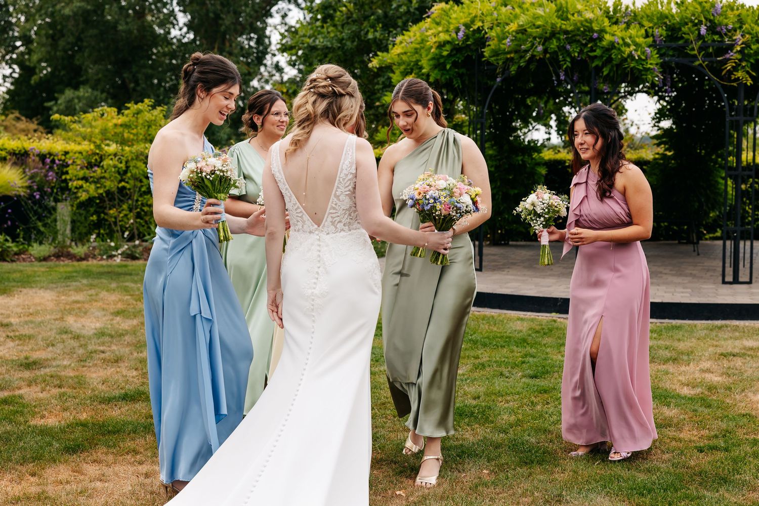 Bride in backless lace gown surrounded by bridesmaids in blue, green, and pink pastel dresses outdoors.